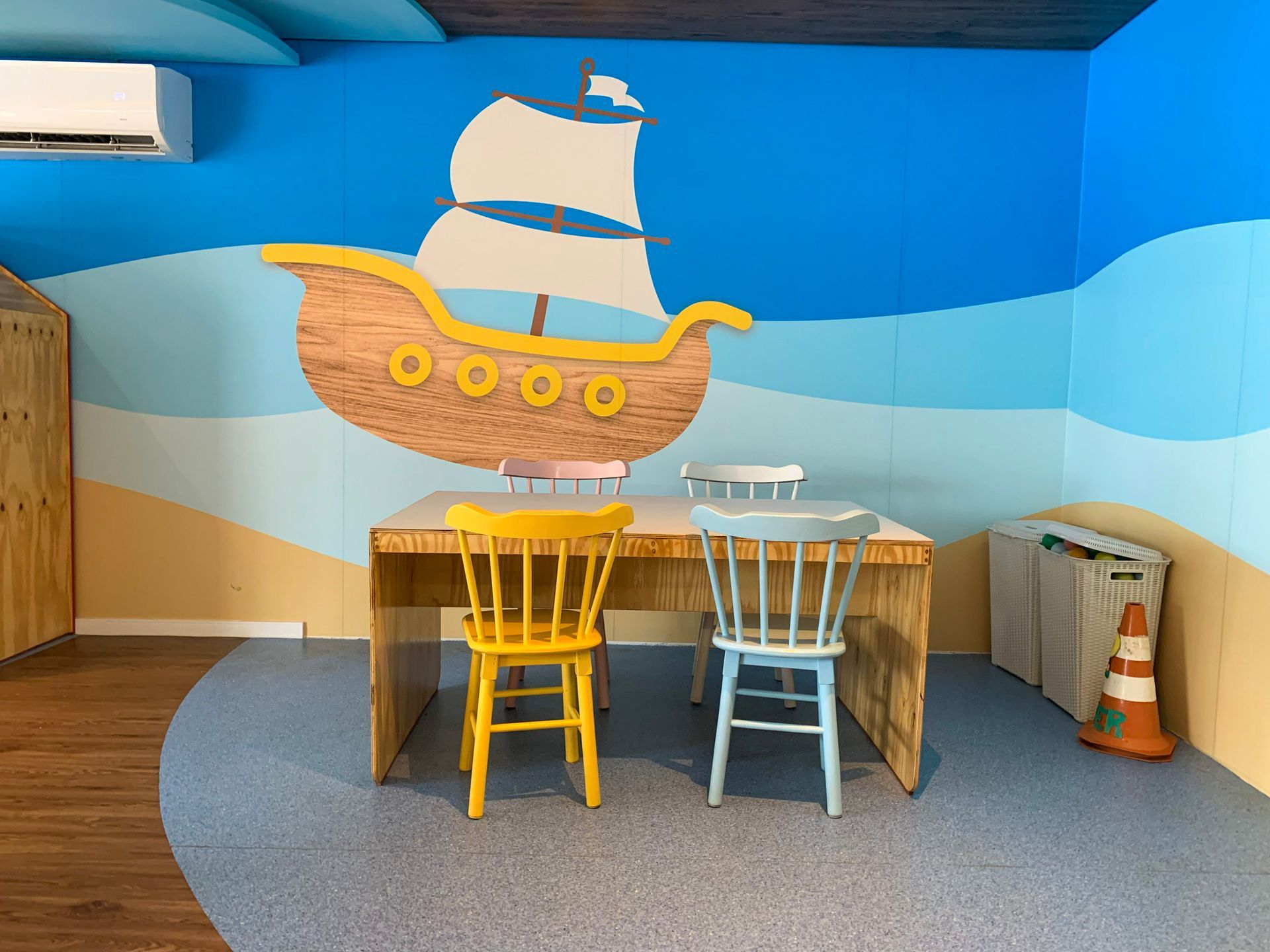 Quarto infantil com um mural pintado de um navio pirata, paredes azuis, mesinhas e cadeiras coloridas.