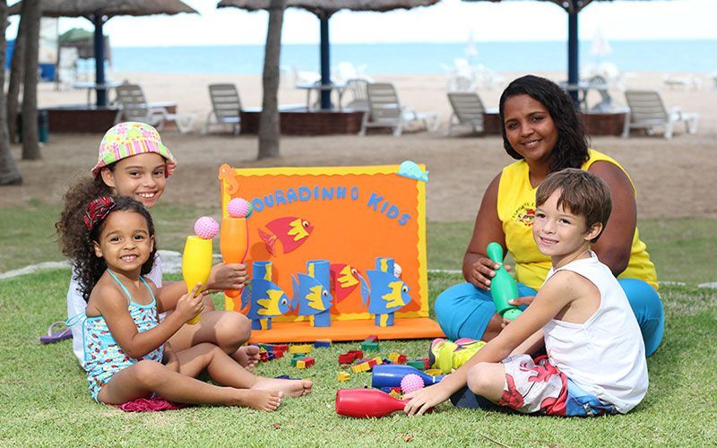 Crianças e um adulto sentam-se na grama em uma praia, brincando com uma placa colorida e brinquedos.
