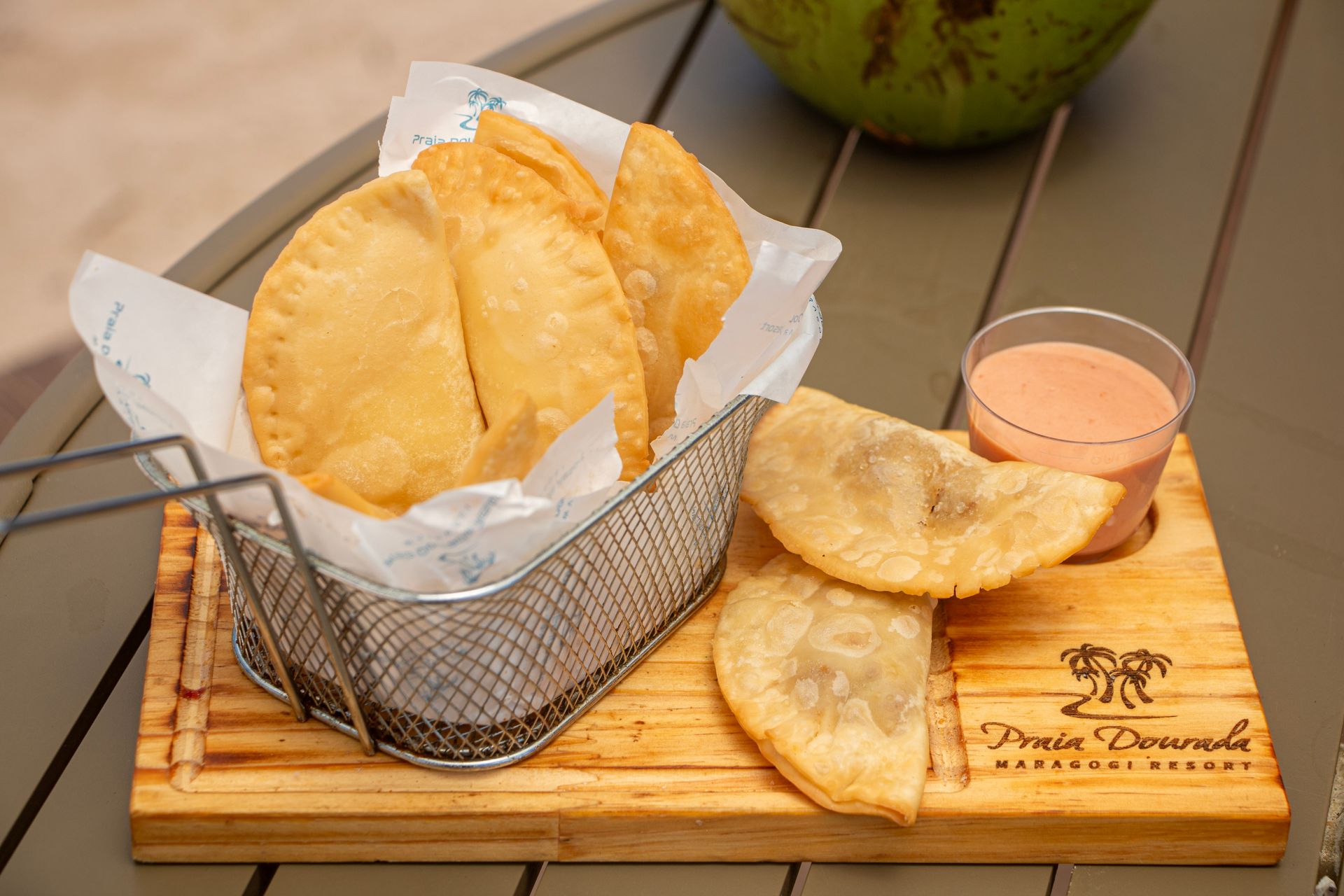 Empanadas fritas em uma cesta de arame e em uma tábua de madeira com molho para mergulhar, servidas sobre uma mesa.