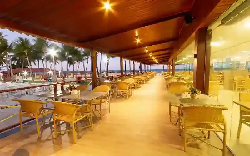 Terraço coberto de um restaurante à beira-mar com mesas e cadeiras vazias, iluminação aconchegante e palmeiras.
