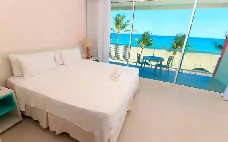 Quarto minimalista à beira-mar com cama branca e vista deslumbrante para o oceano através de grandes janelas.