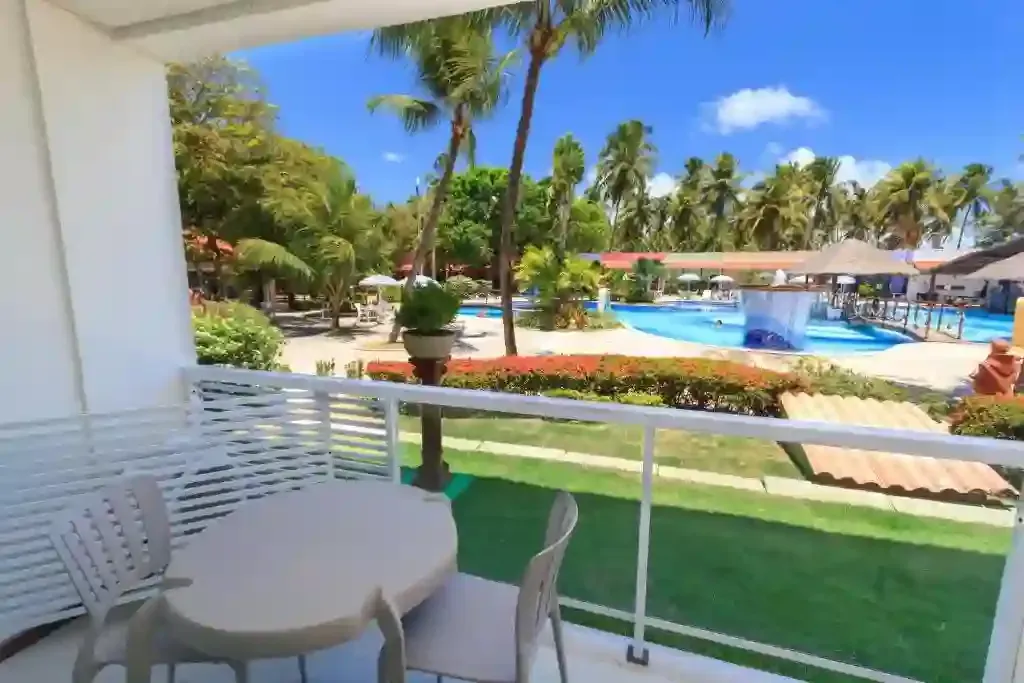 Varanda com mesa de pátio branca com vista para a piscina de um resort tropical, palmeiras e céu azul.