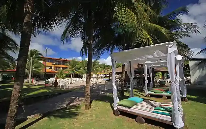 Pátio de resort tropical com palmeiras, cabanas brancas e espreguiçadeiras à beira da piscina sob um céu azul.