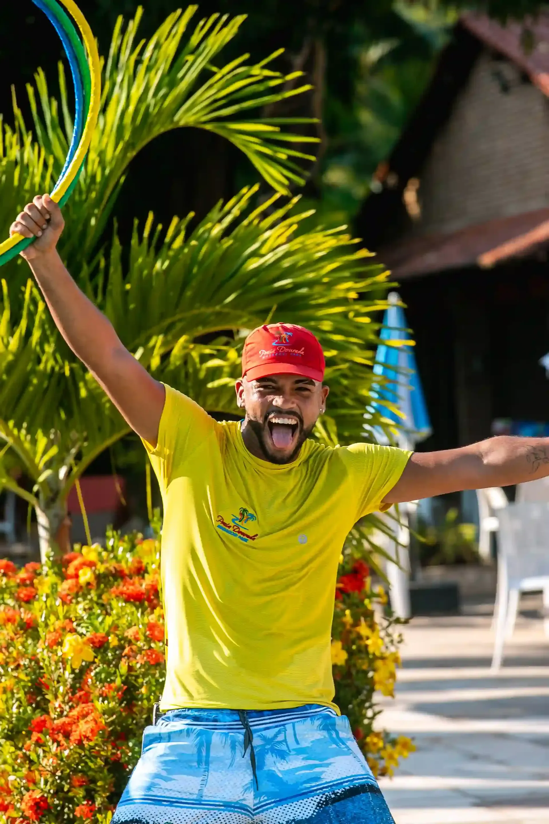 Homem de camisa amarela e boné vermelho dançando ao ar livre com os braços erguidos, sorrindo em meio a plantas tropicais.