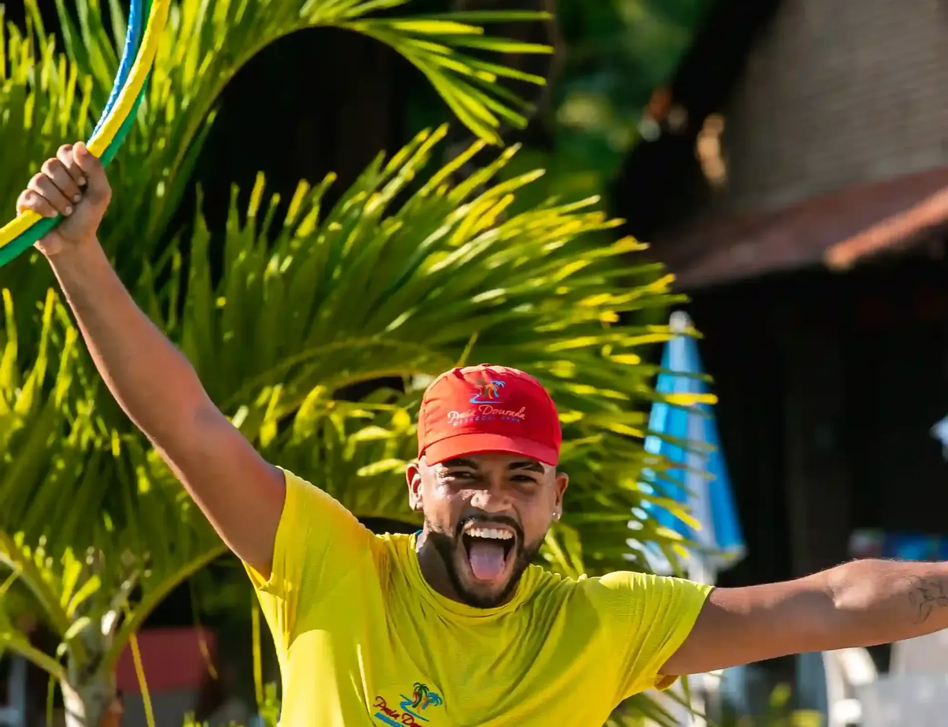 Homem de boné vermelho e camisa amarela comemorando com os braços erguidos ao ar livre em meio a plantas tropicais.
