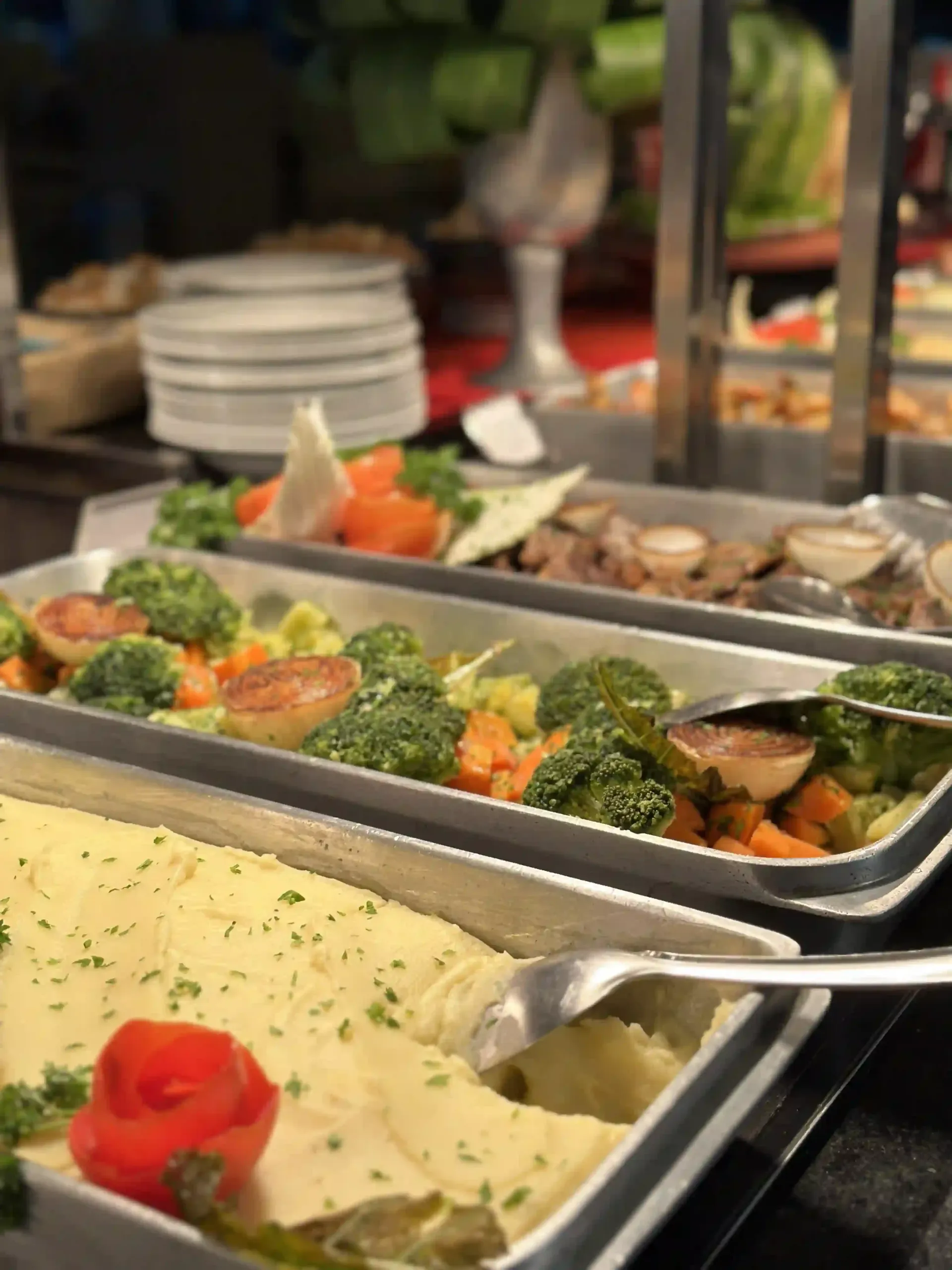 Buffet com bandejas de legumes variados, purê de batatas e pratos individuais em um ambiente de restaurante.