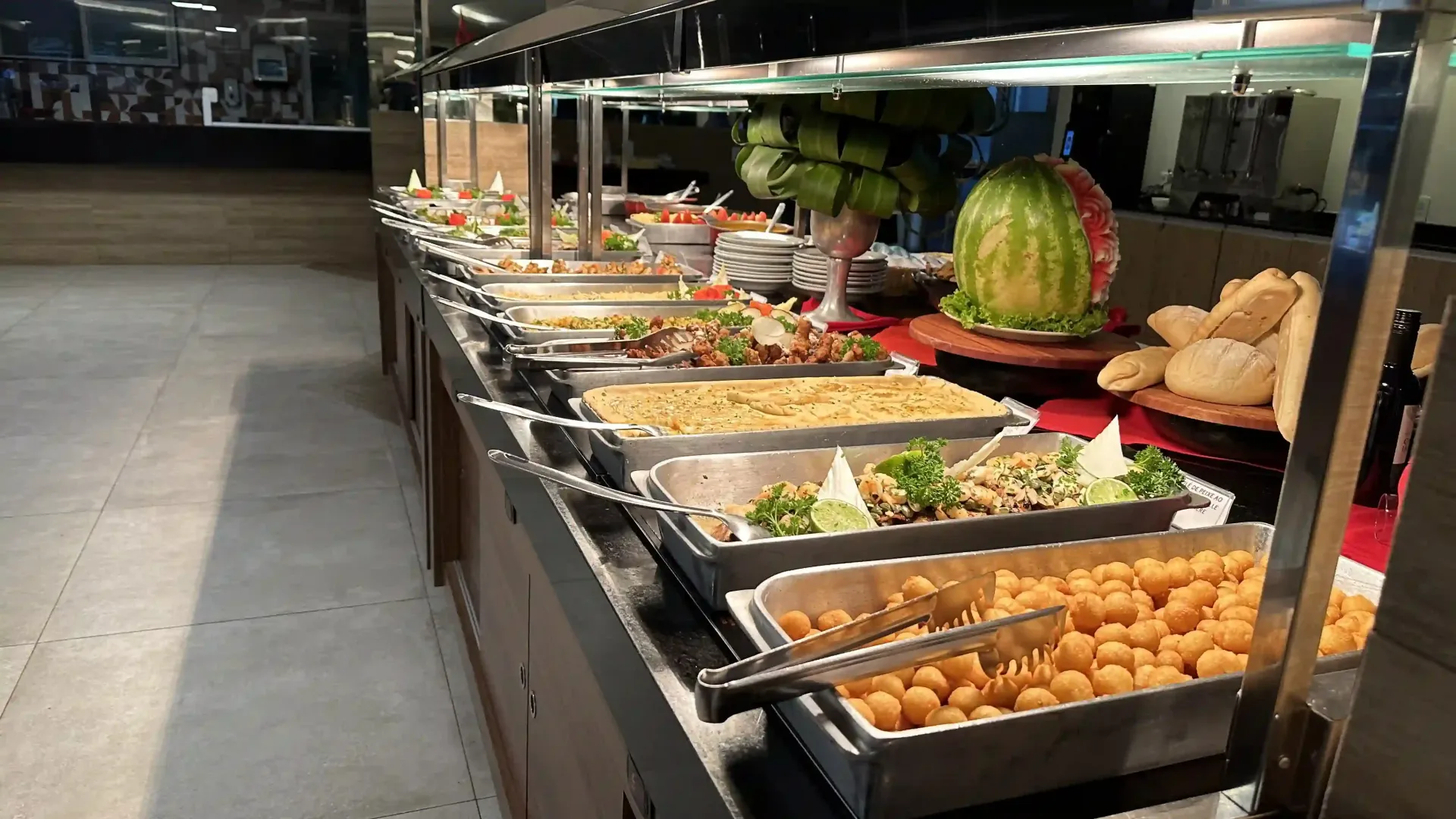 Mesa de buffet com bandejas de comida variadas, pão, frutas e uma melancia em um restaurante com iluminação suave.