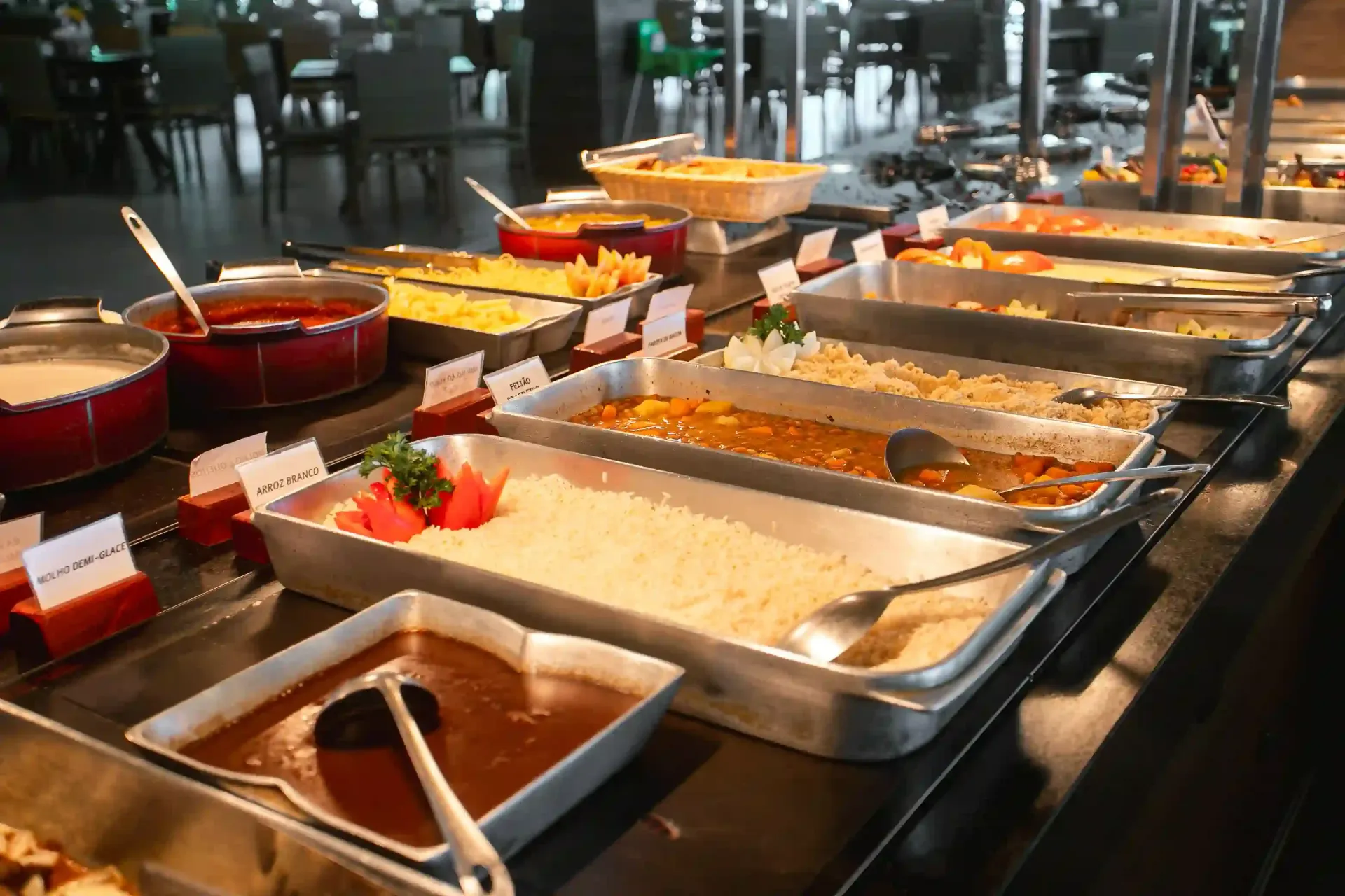 Buffet com bandejas de aço inoxidável contendo diversos pratos e molhos em um ambiente de restaurante.
