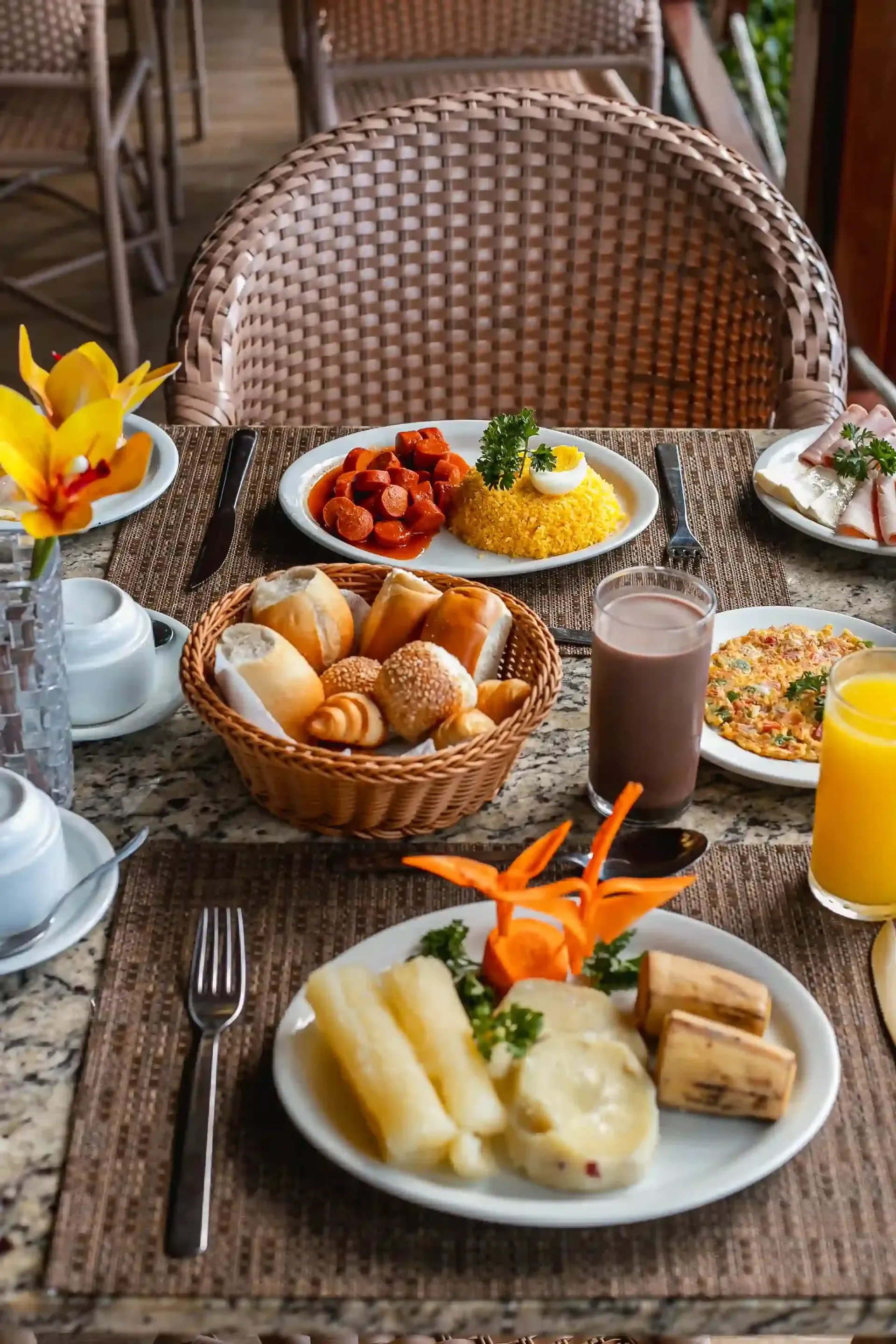 Café da manhã servido em uma mesa de vime com frutas, pão, suco e café ao lado de uma cadeira.