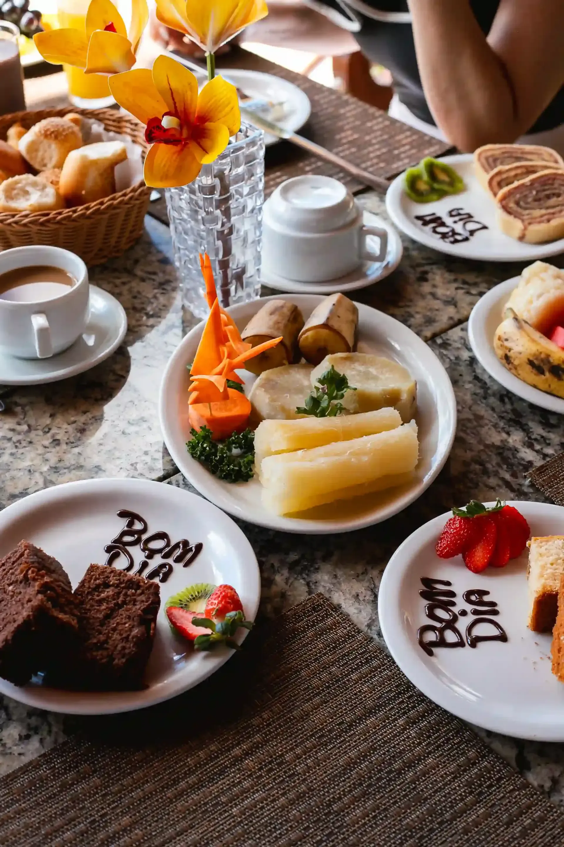 Mesa de brunch com chá, café, doces, frutas e pão em pratos brancos ao redor de um vaso de flores.
