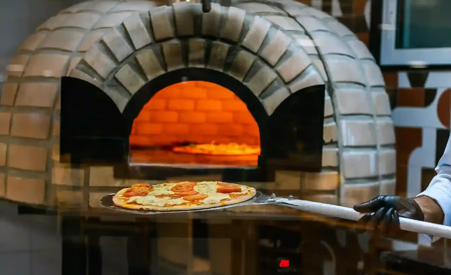 Pizza assando em forno de tijolos, com uma pizza em uma pá de pizza em primeiro plano.