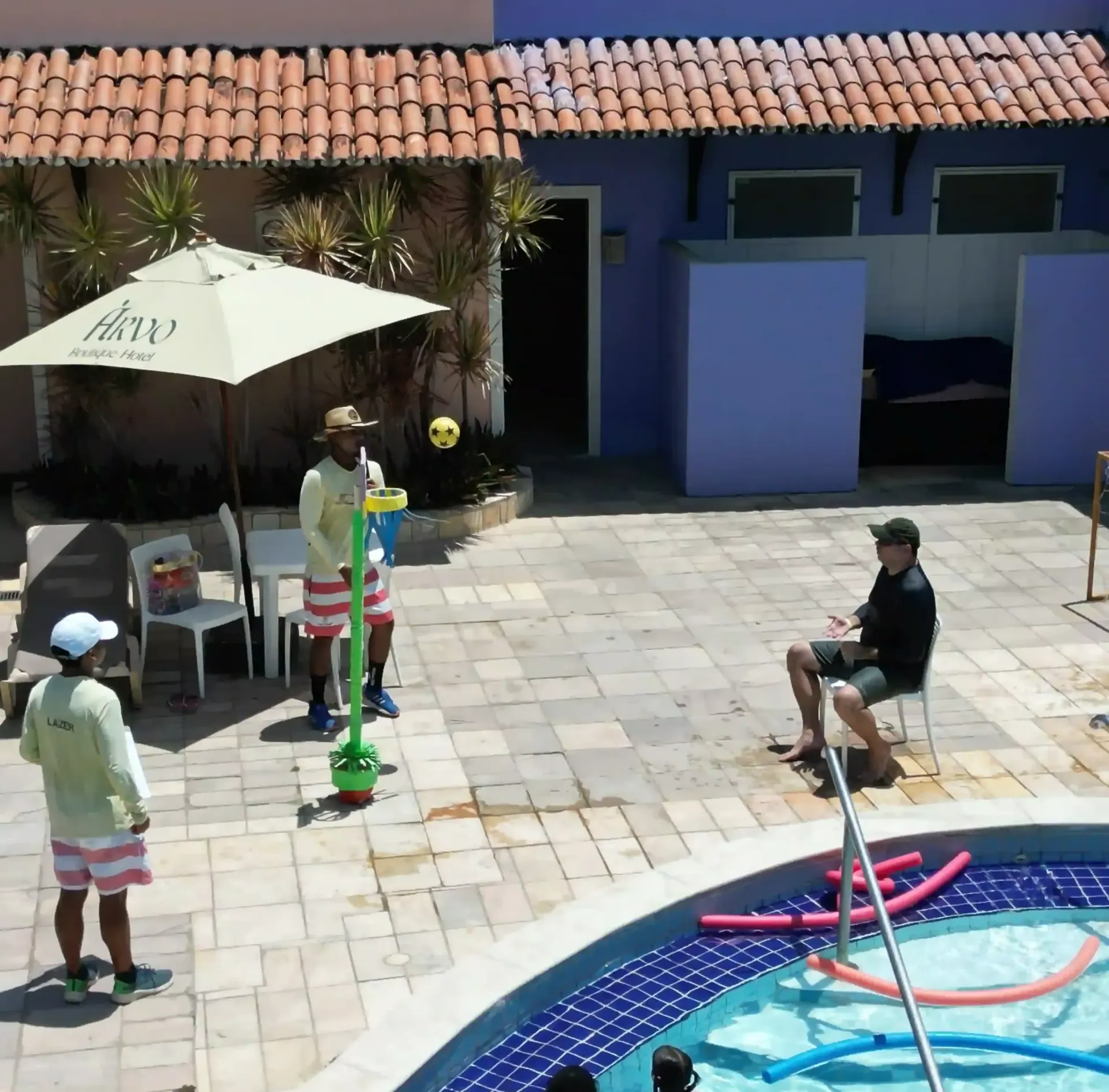 Pessoas em um pátio à beira da piscina perto de um prédio azul, uma delas sentada e as outras em pé sob um guarda-sol.
