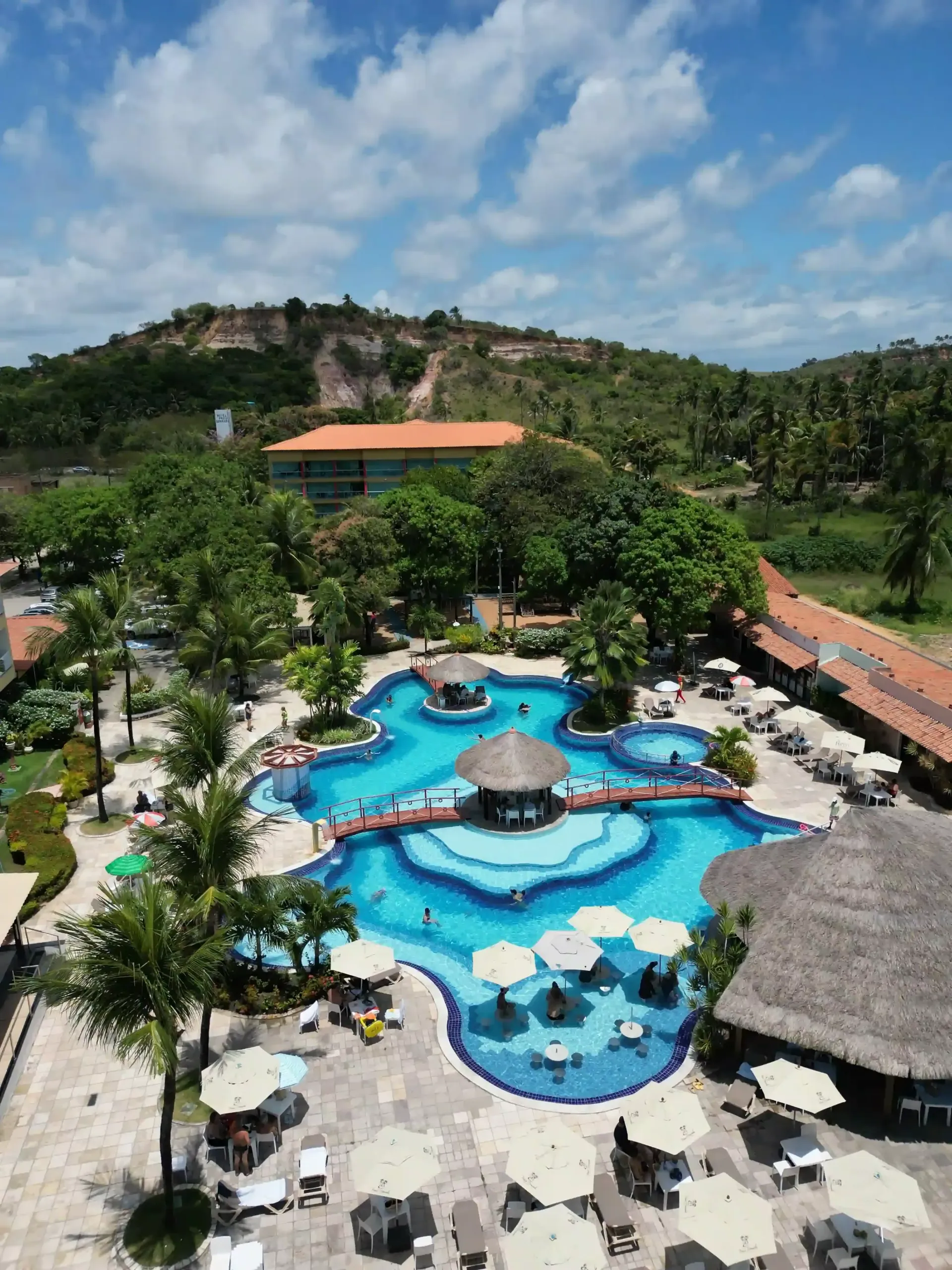Piscina do resort com água azul, palmeiras, espreguiçadeiras e cabanas de palha sob um céu ensolarado.