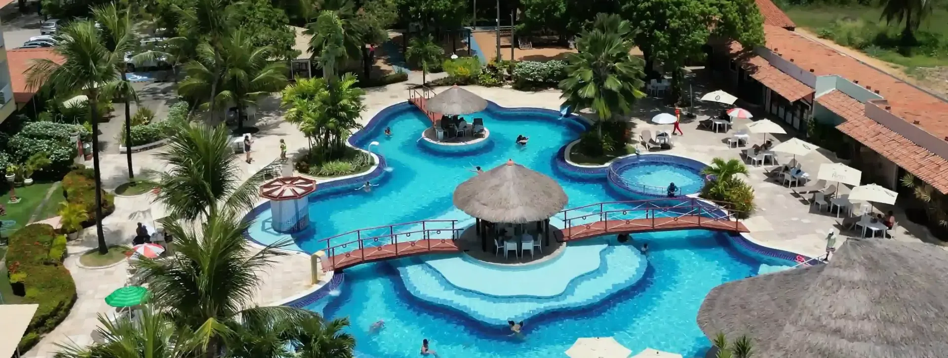 Vista aérea de um complexo de piscinas de um resort com piscinas azuis, pontes curvas e um gazebo com teto de palha.