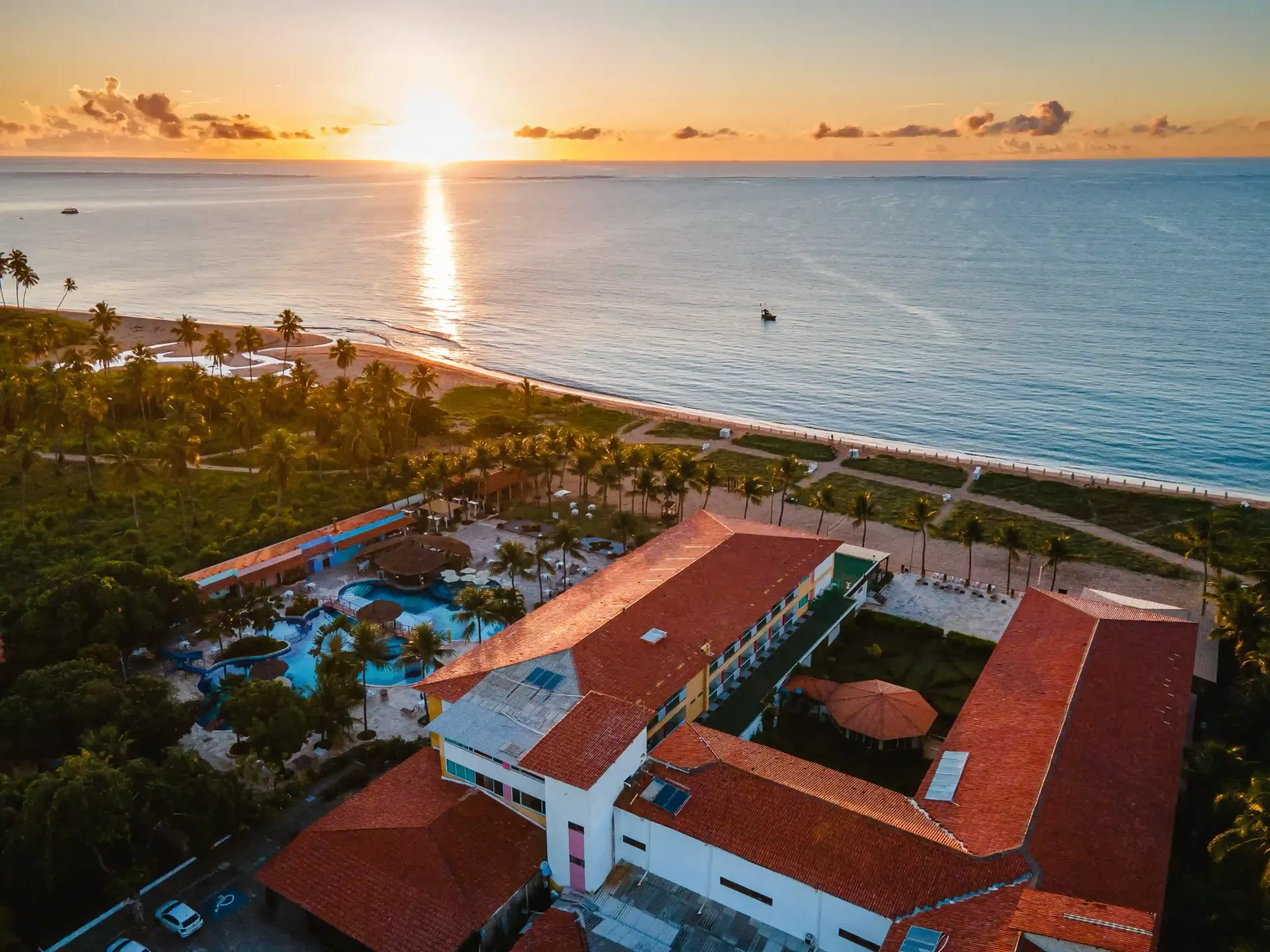 Pôr do sol sobre um resort costeiro com edifícios de telhados vermelhos e ondas do oceano.
