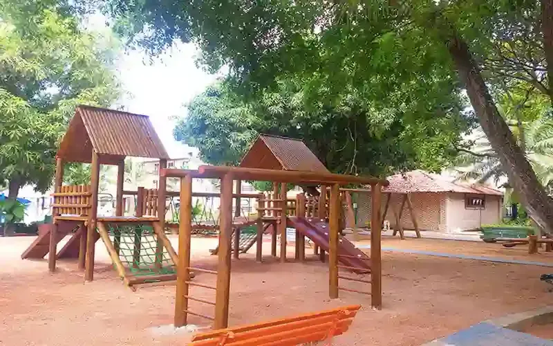 Estruturas de madeira para parque infantil com escorregadores sob árvores frondosas em uma área de parque arenoso.