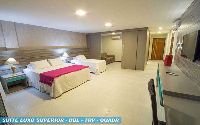 Suíte de hotel moderna com duas camas, sofá, escrivaninha e ampla janela panorâmica.