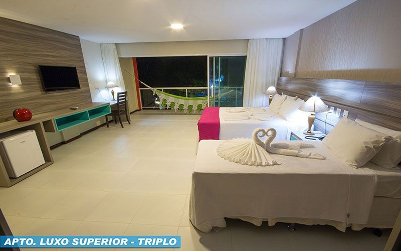 Quarto de hotel com duas camas, TV, escrivaninha e varanda com vista para a vegetação à noite.