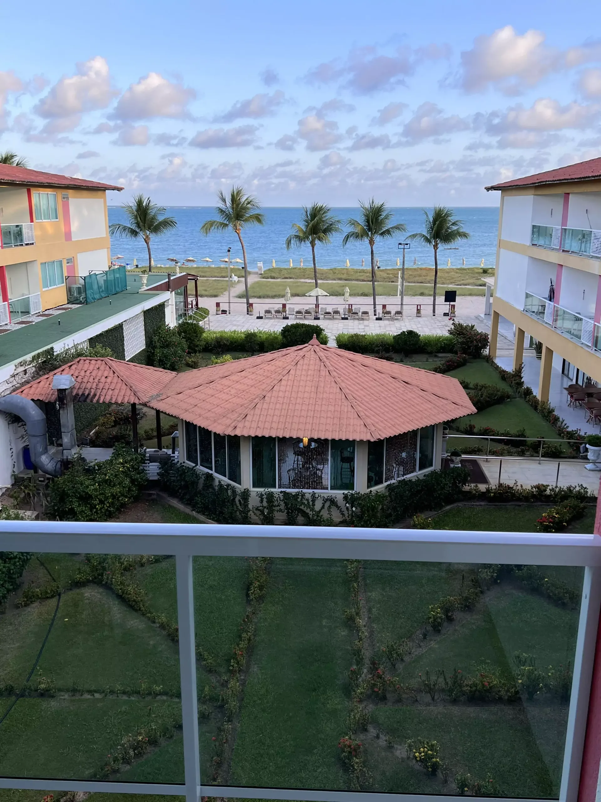 Pátio do resort com um pavilhão de telhado vermelho, palmeiras e vista para o mar entre dois edifícios em tons pastel.