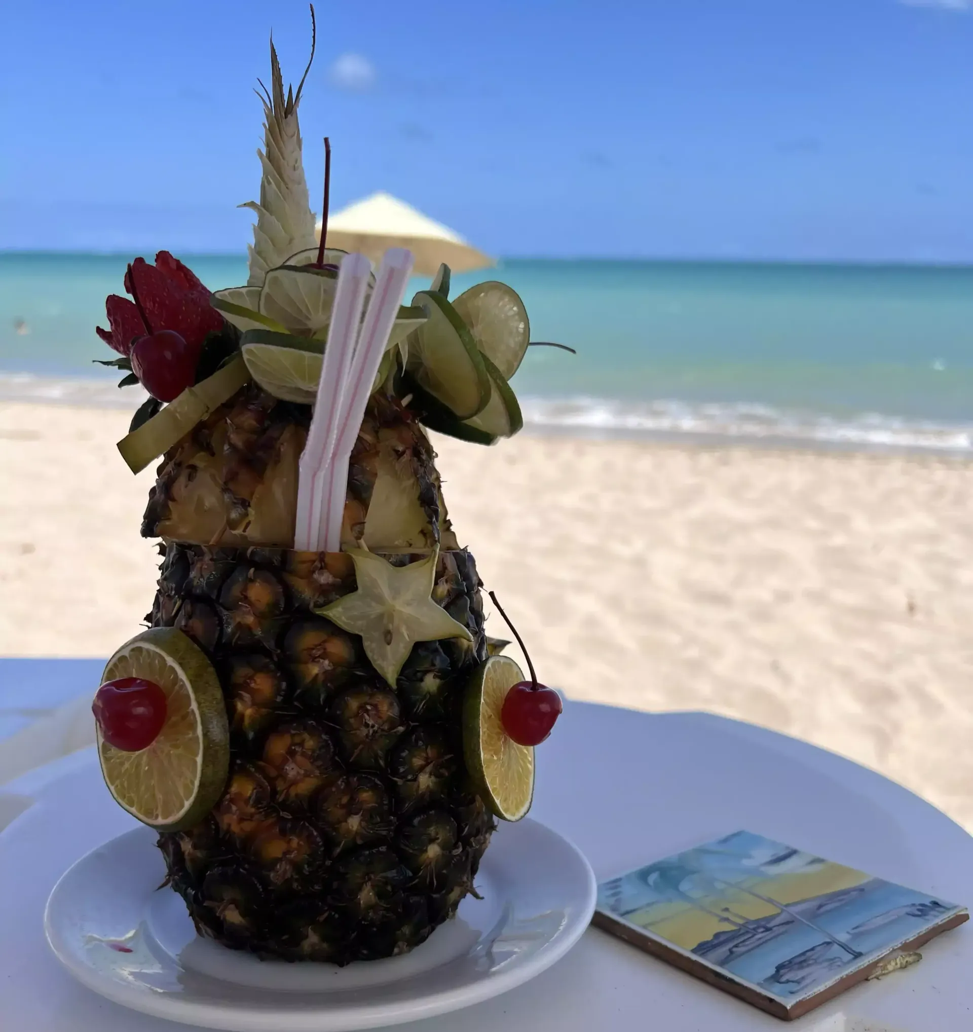 Bebida tiki de abacaxi com decoração de frutas em uma mesa de praia, com o oceano e a areia ao fundo.