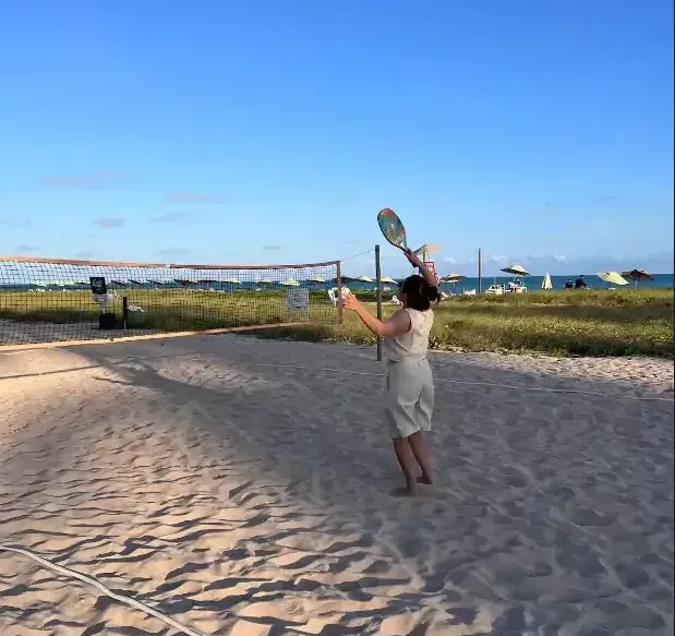 Pessoa jogando tênis em uma quadra de areia perto da praia sob um céu azul claro.