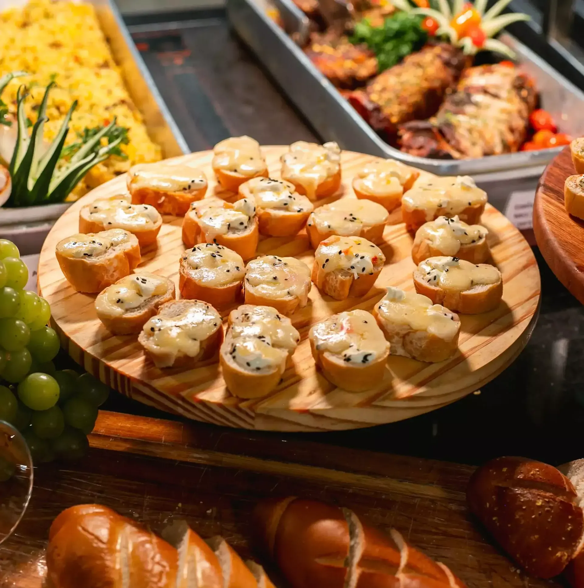 Canapés em uma bandeja de madeira em um bufê, com arroz, carne grelhada e frutas ao fundo.