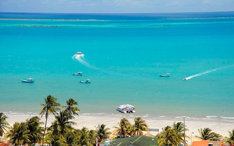 Praia tropical com água turquesa, palmeiras e vários barcos perto da costa.