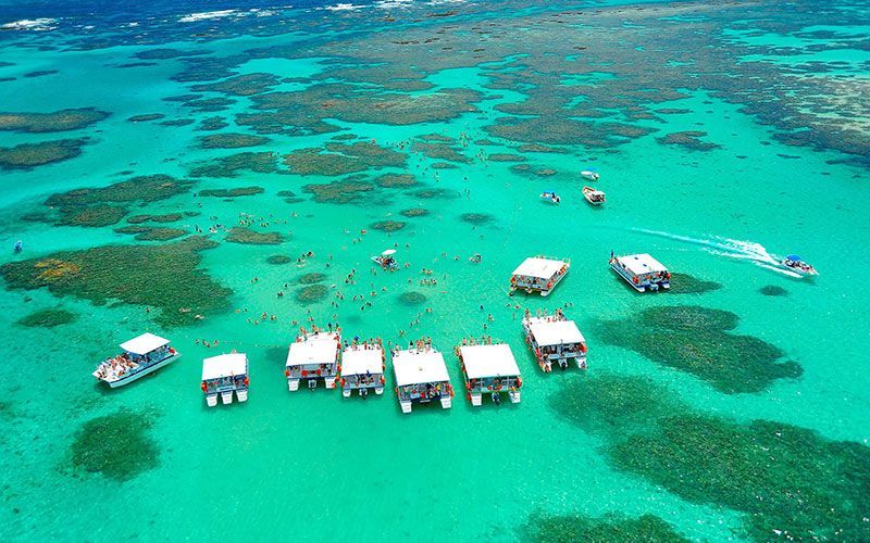 Vista aérea de barcos brancos em águas rasas de cor turquesa com manchas de coral e um banco de areia.