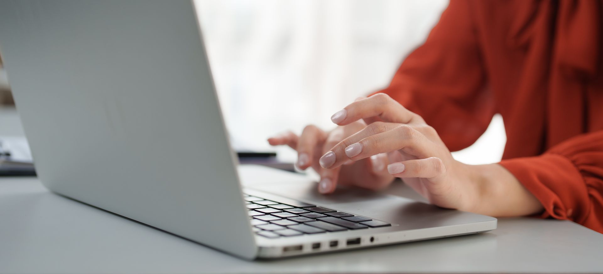 A person in a rust-colored top typing on a silver laptop.