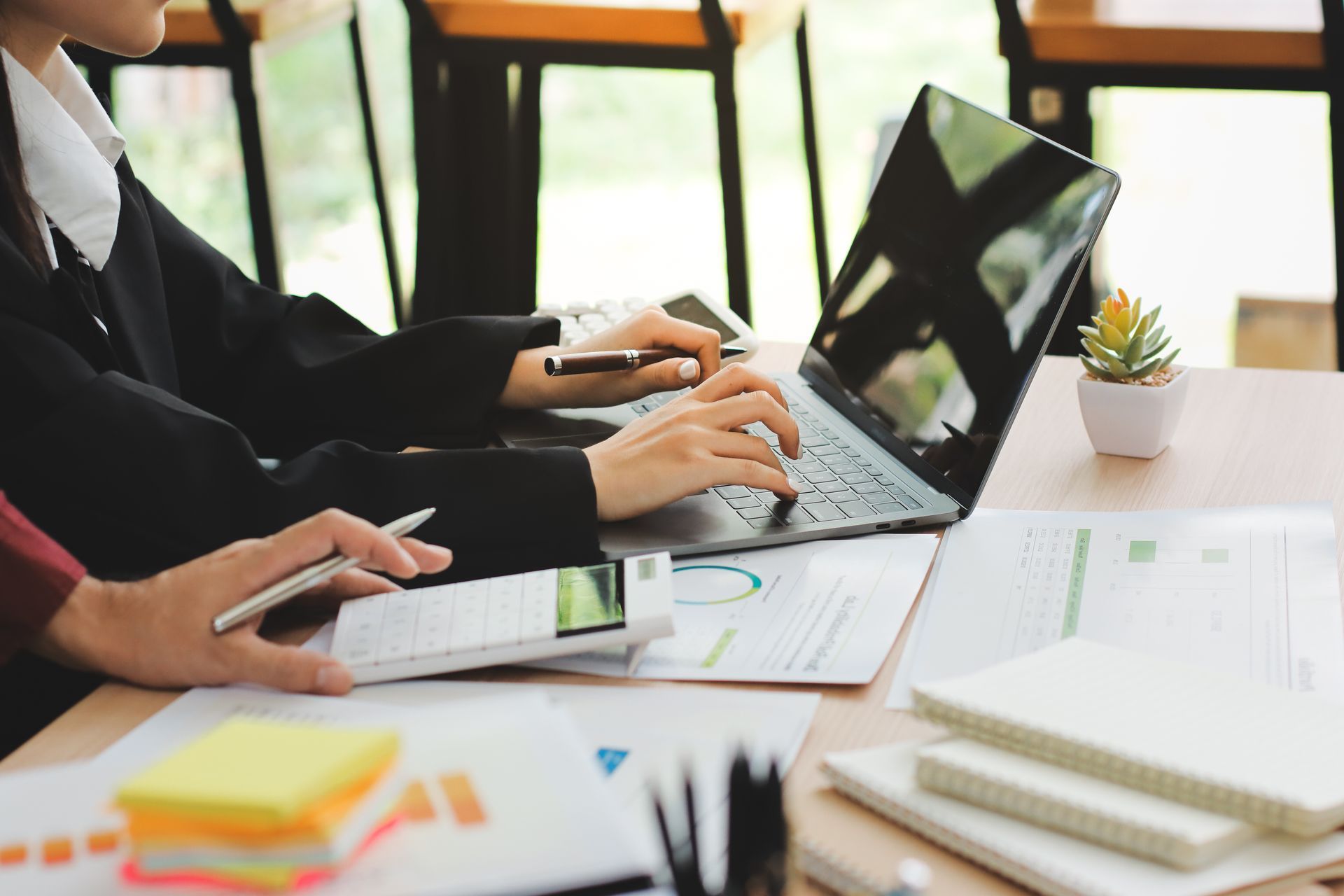 People working on laptops at a bright office table with notebooks, sticky notes, and a small plant