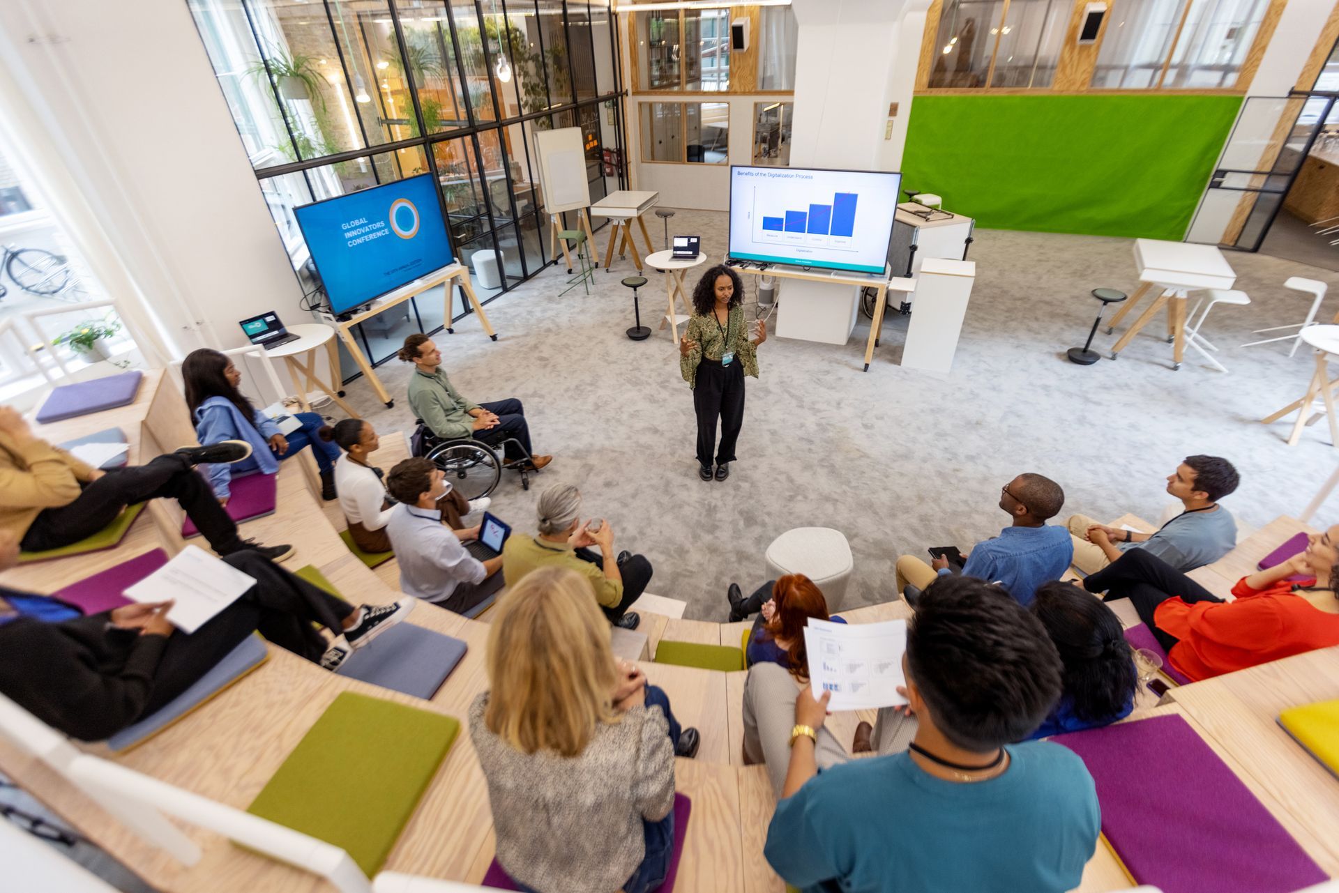 A presenter speaks to a diverse group seated on stepped wooden benches in a modern, bright, open-plan office space.