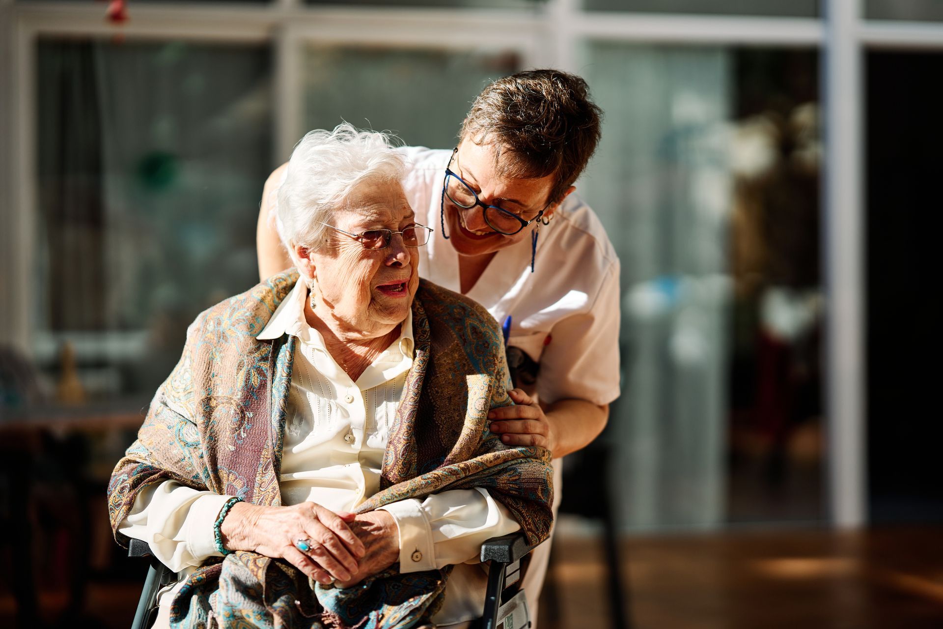 A caregiver with glasses smiles warmly while placing a hand on the shoulder of an individual sitting in a wheelchair.