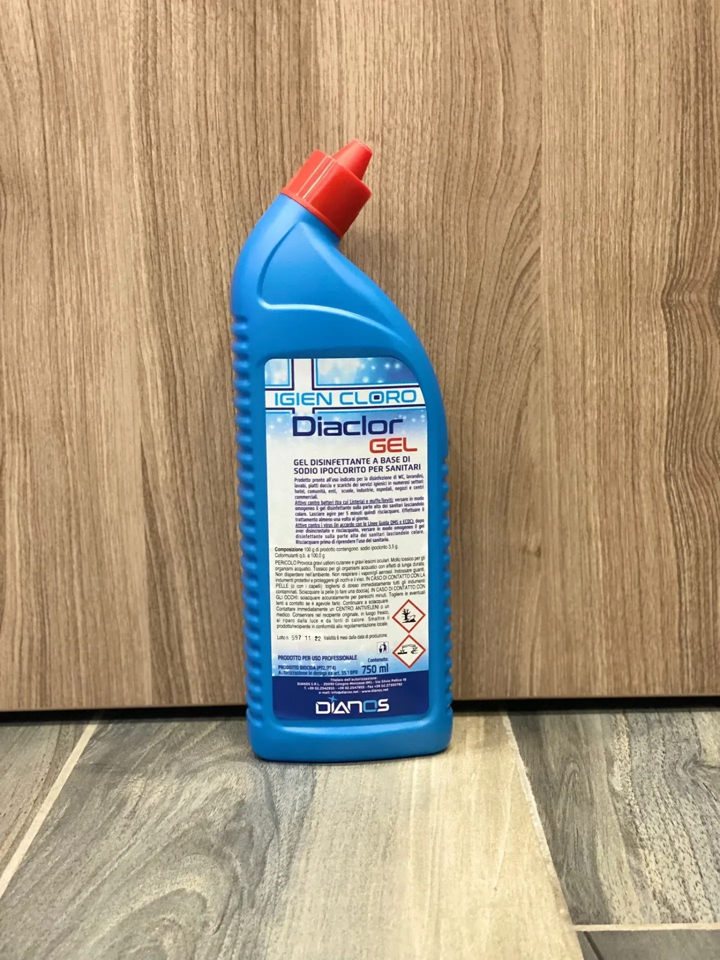 Flacone blu per detergente per WC con tappo rosso, appoggiato su un pavimento in legno, con uno sfondo di pannelli in legno.
