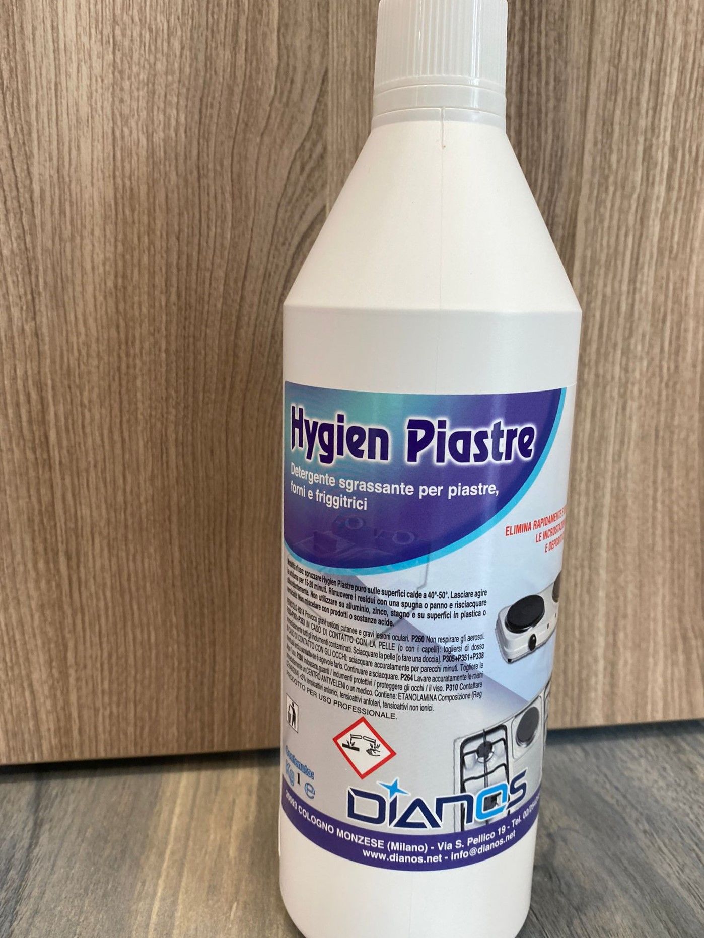 Flacone bianco di detergente Hygien Piastre con etichetta viola su un pavimento di legno, accanto a un mobiletto di legno.