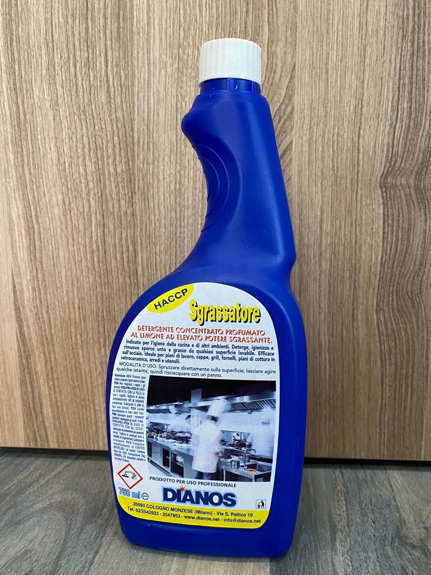 Flacone spray detergente blu con tappo ed etichetta bianchi, appoggiato su una superficie di legno.