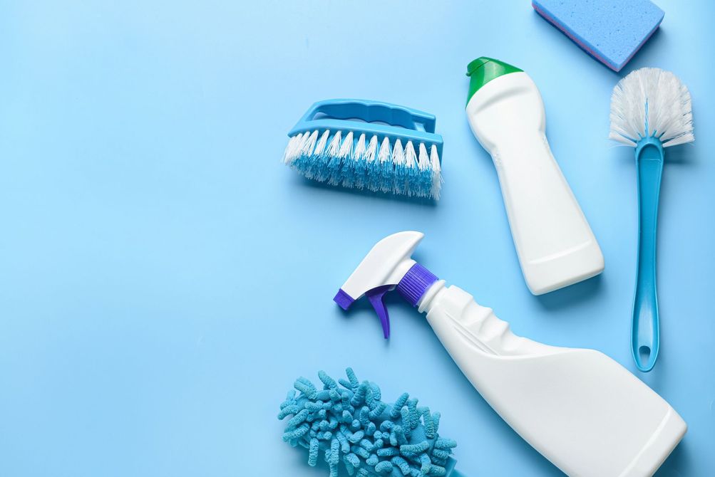 Una persona con tuta protettiva bianca e respiratore tiene in mano uno spruzzatore e un serbatoio di disinfettante in un ambiente interno.