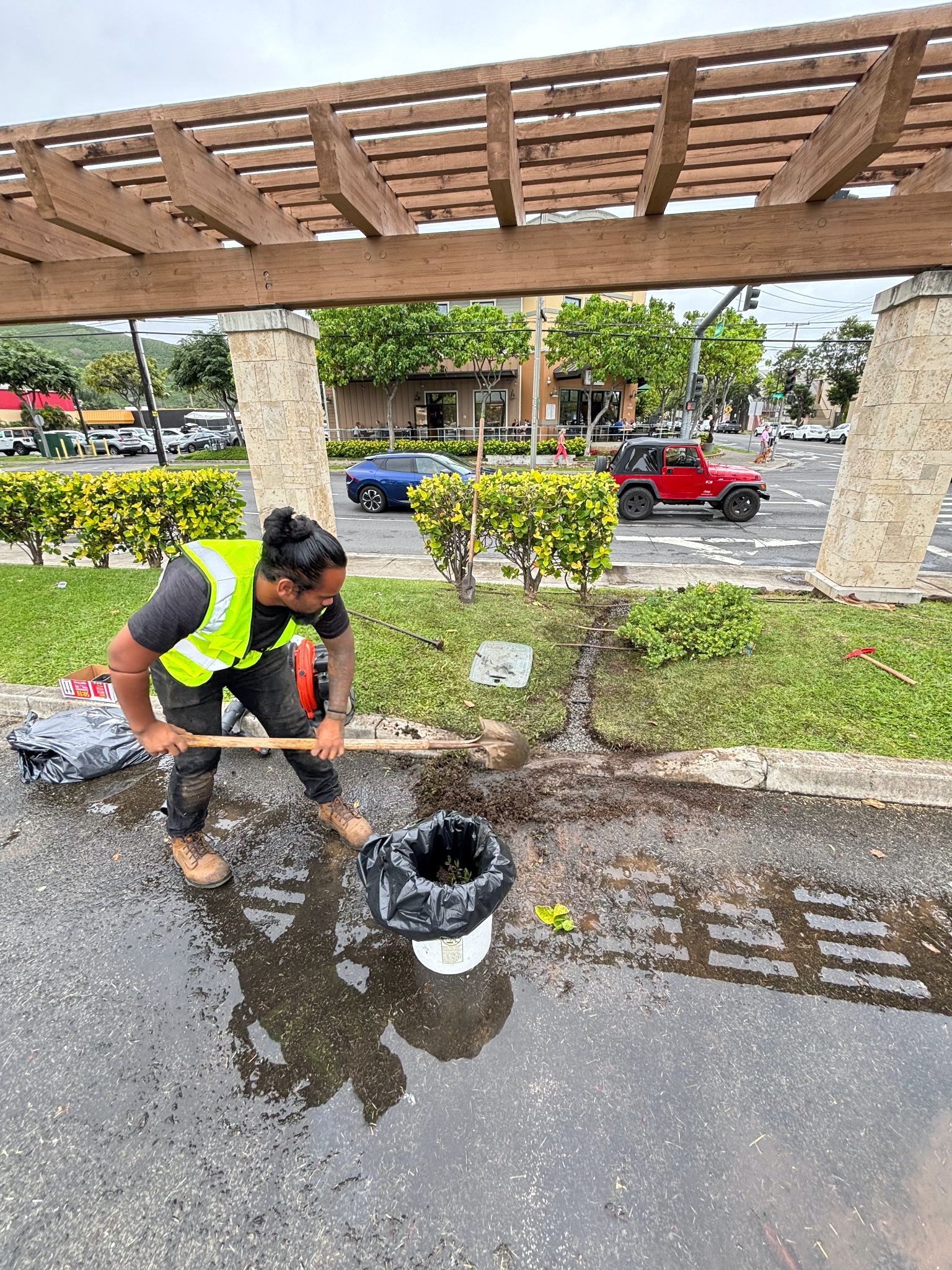 A worker in a high-visibility vest uses a hoe to clear debris near a landscaped area during a rainy day.