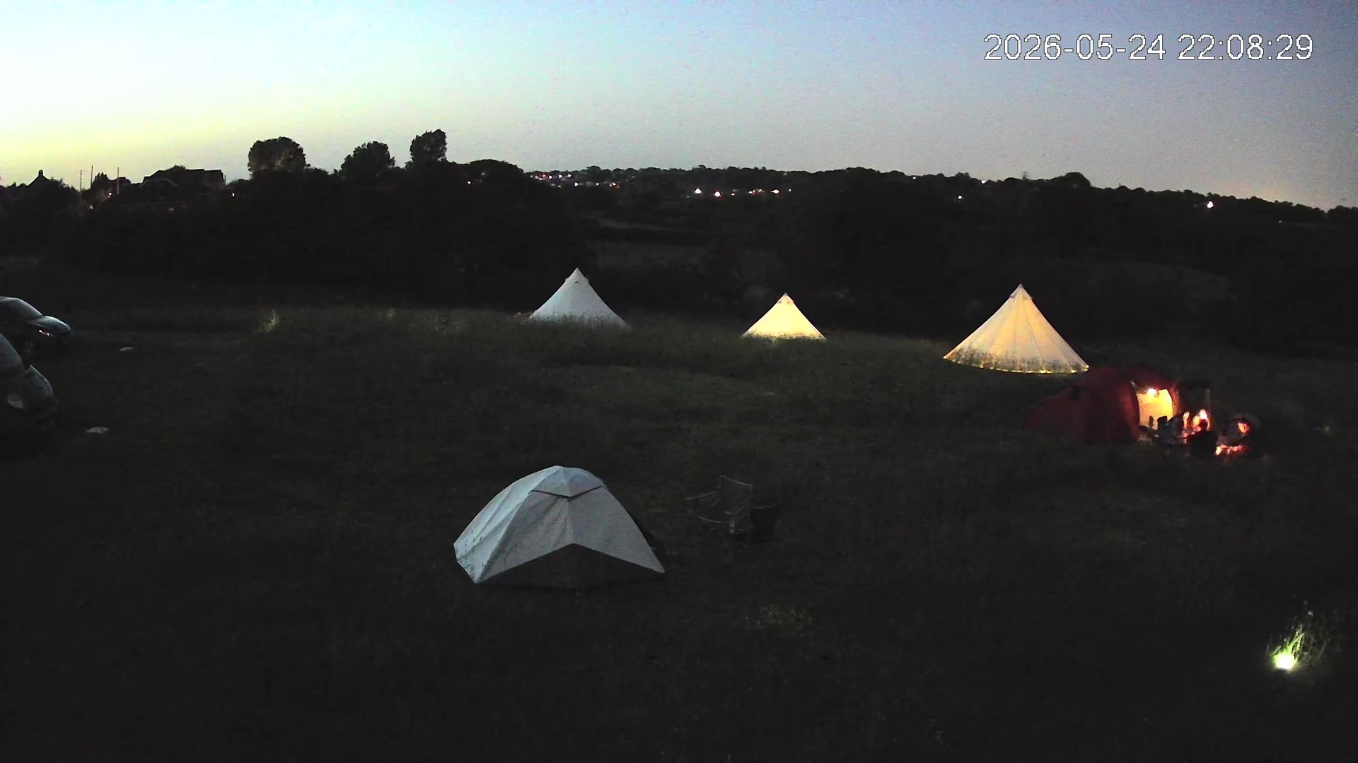 Dusk campsite with several glowing tents on a dark field and a small campfire at right