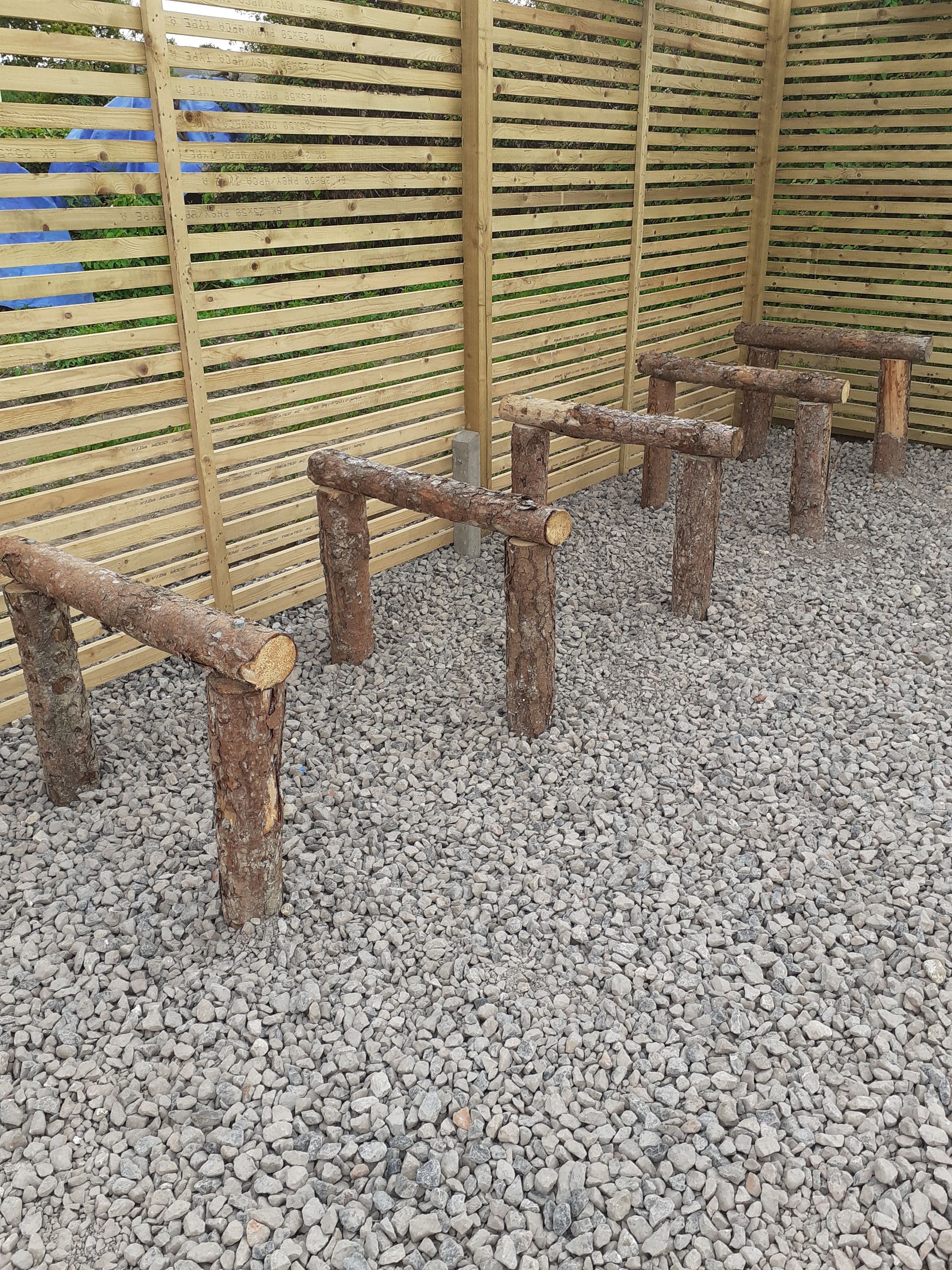 Wooden log benches on gravel; next to a slatted wood fence.