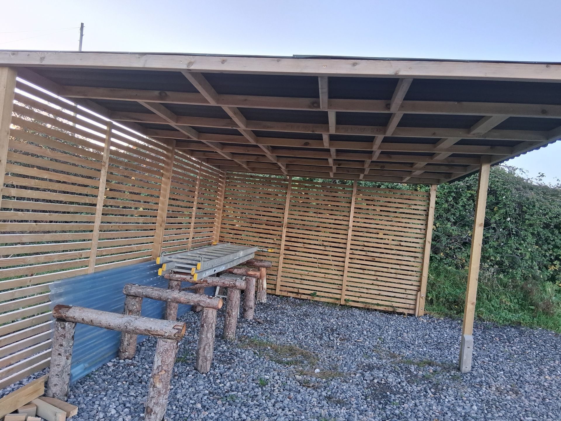 Wooden bike shelter with slatted walls and roof, gravel floor. Blue sky in background.