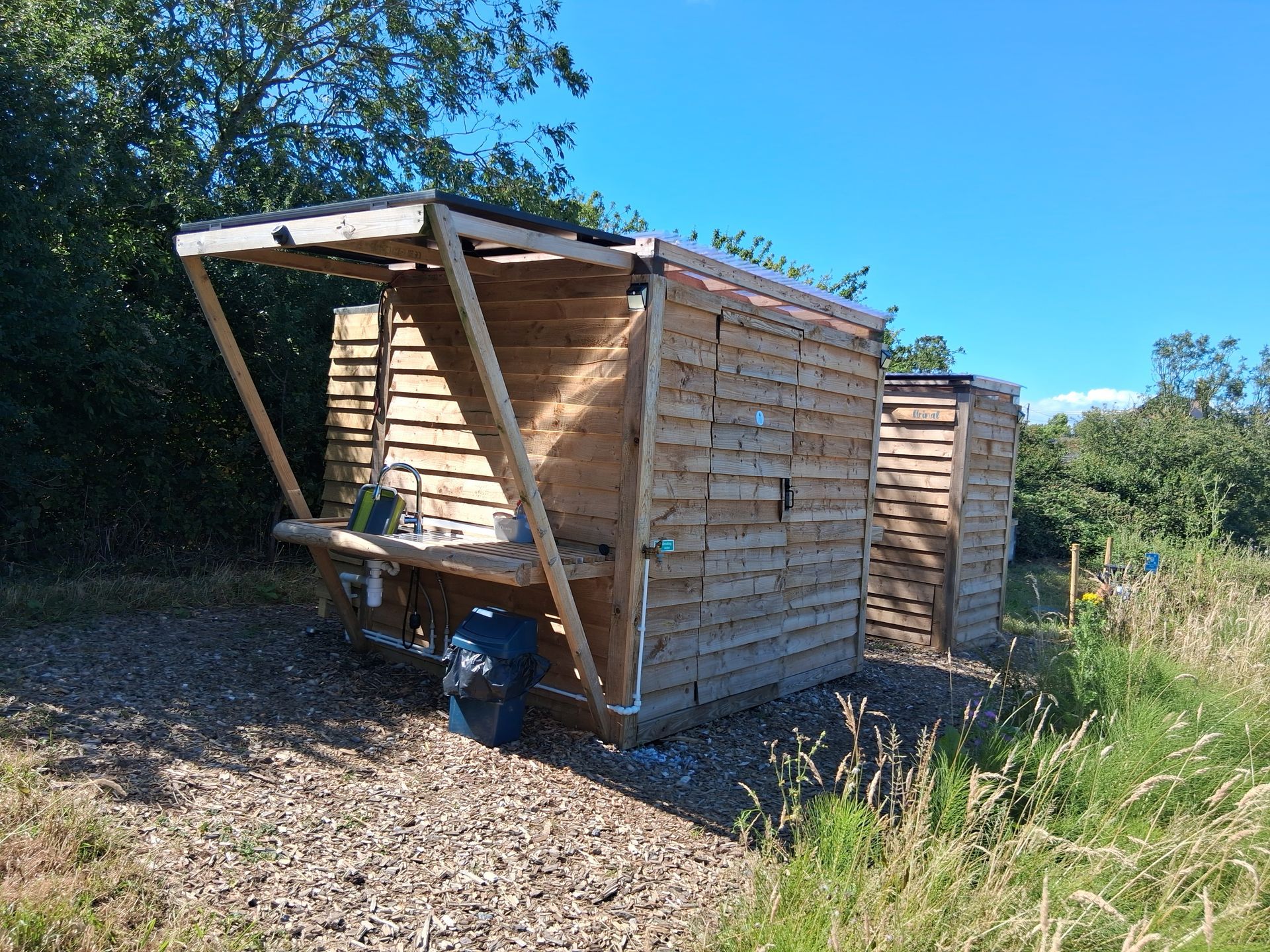 Wooden shed with a slanted roof and open front, sitting in a sunny grassy field under blue sky.
