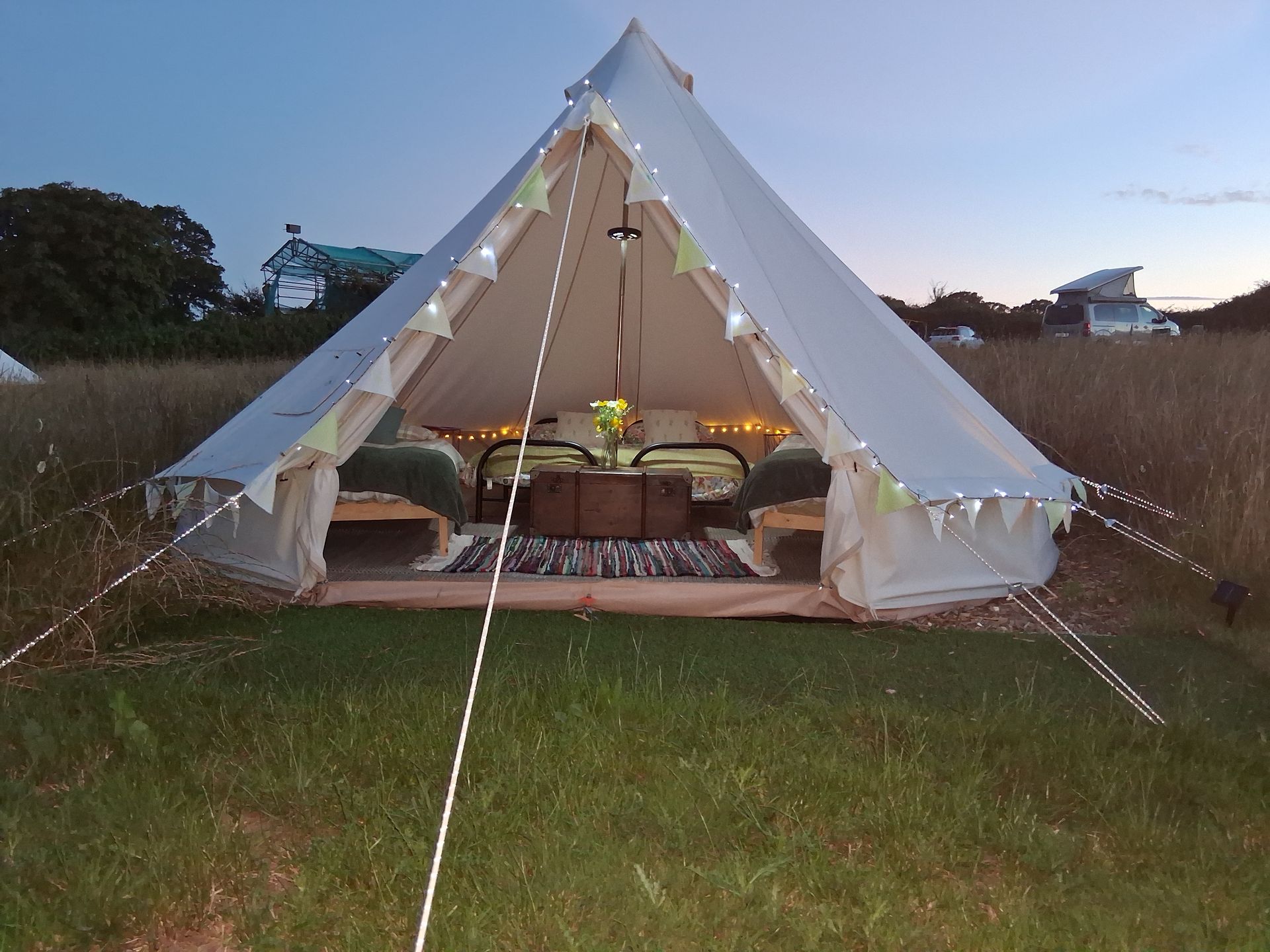 White teepee tent lit at dusk on a grassy field with camping gear inside