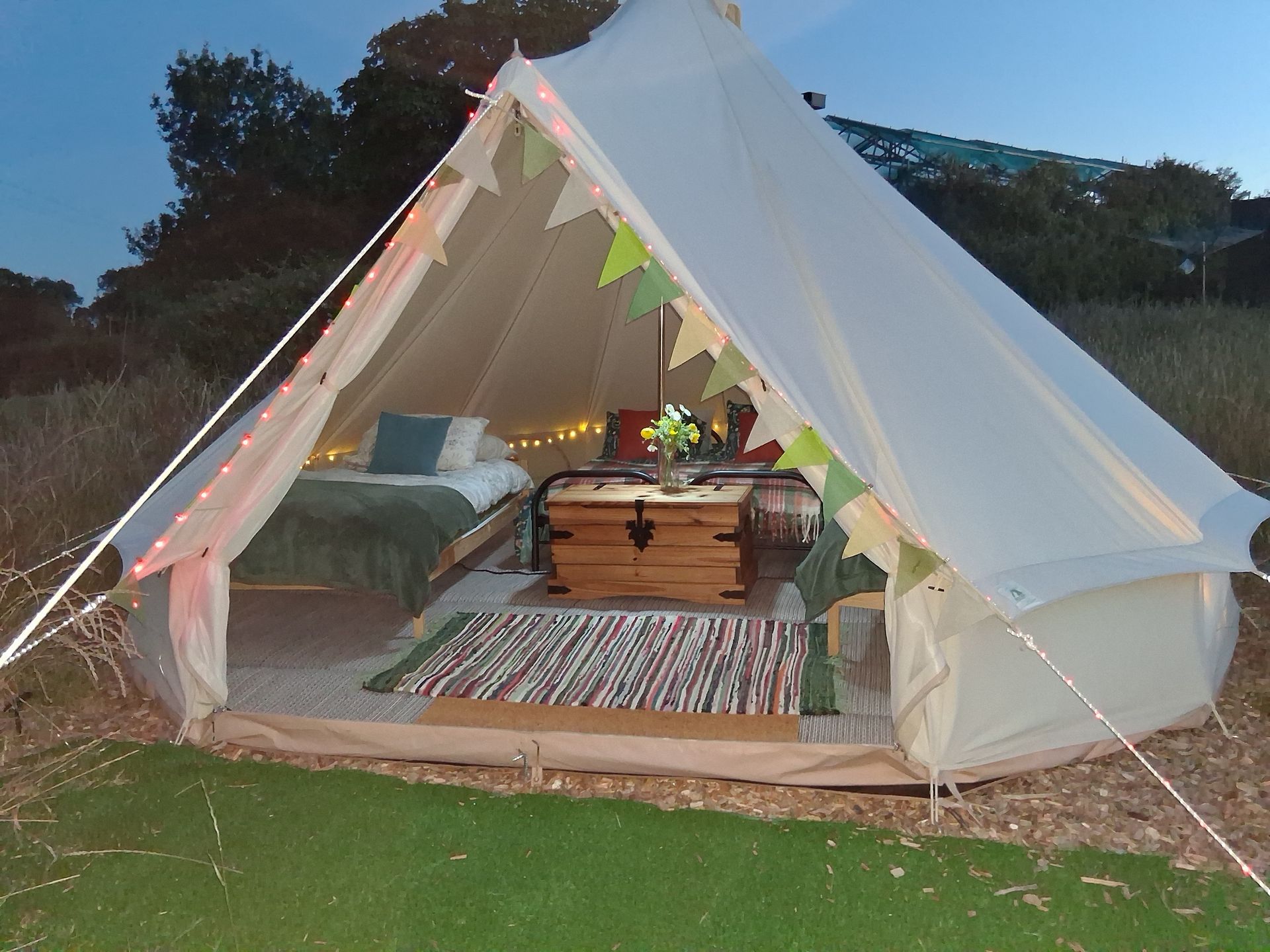 White canvas tent lit at dusk, with cozy interior seating and a wooden chest on a striped rug outdoors