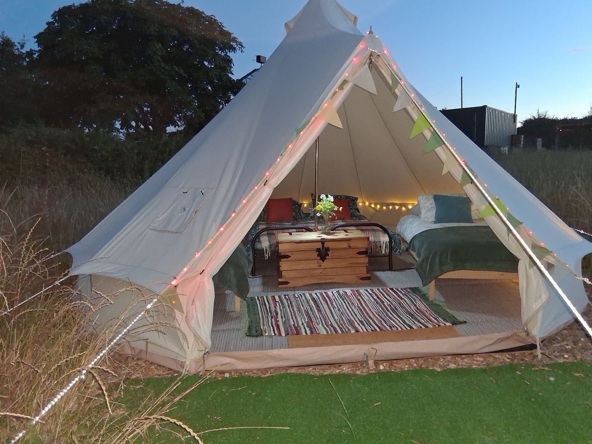 Canvas tent lit at dusk, with wooden bed, rug, and seating inside on grass.