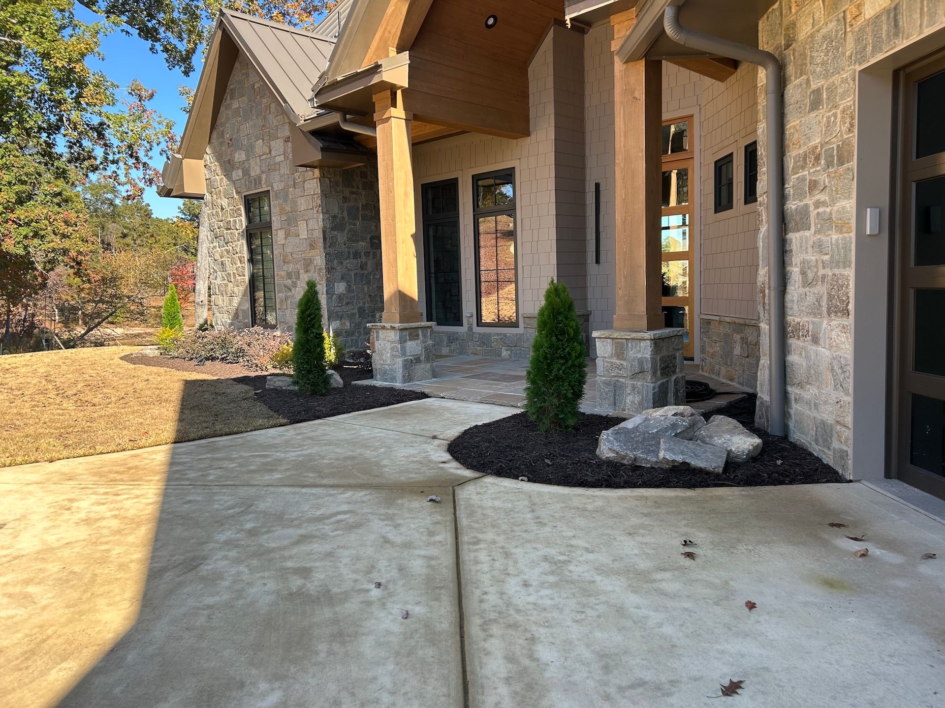 Stone-front house entrance with concrete walkway, black mulch beds, and small shrubs in sunlight