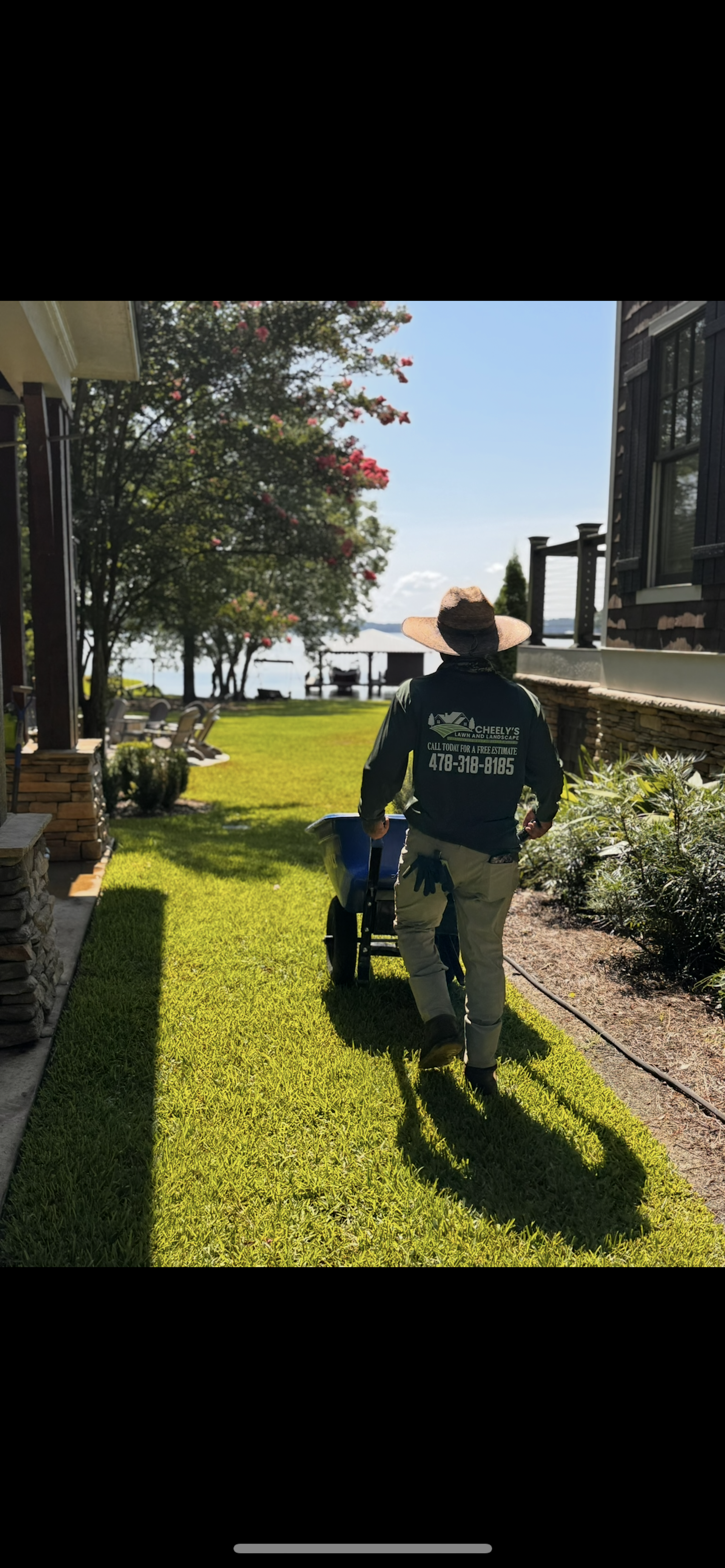 Person pushing a stroller on a sunny grass path beside buildings and water.