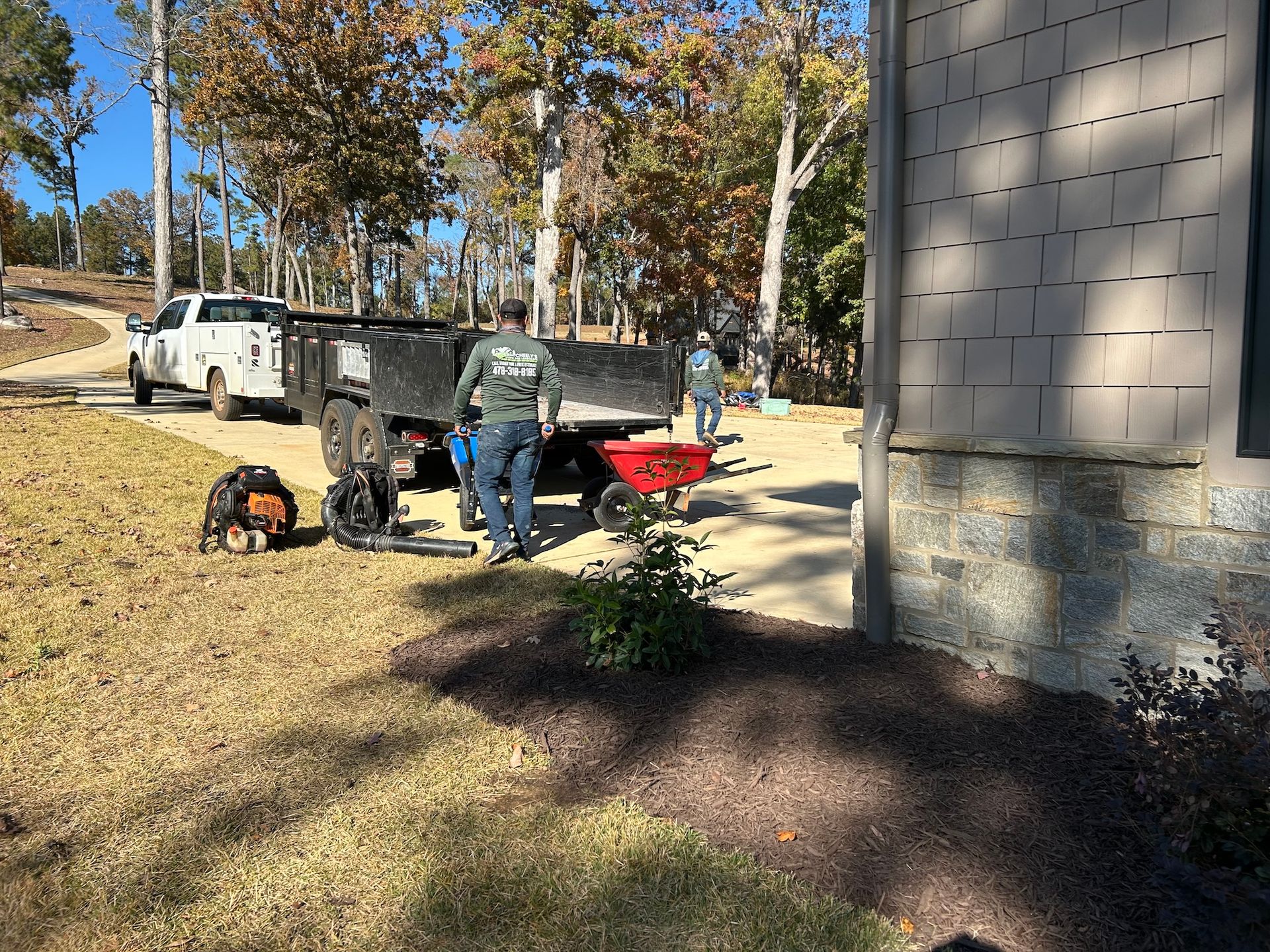 Workers loading equipment beside a white truck and red trailer near a building, with a small dog in the foreground