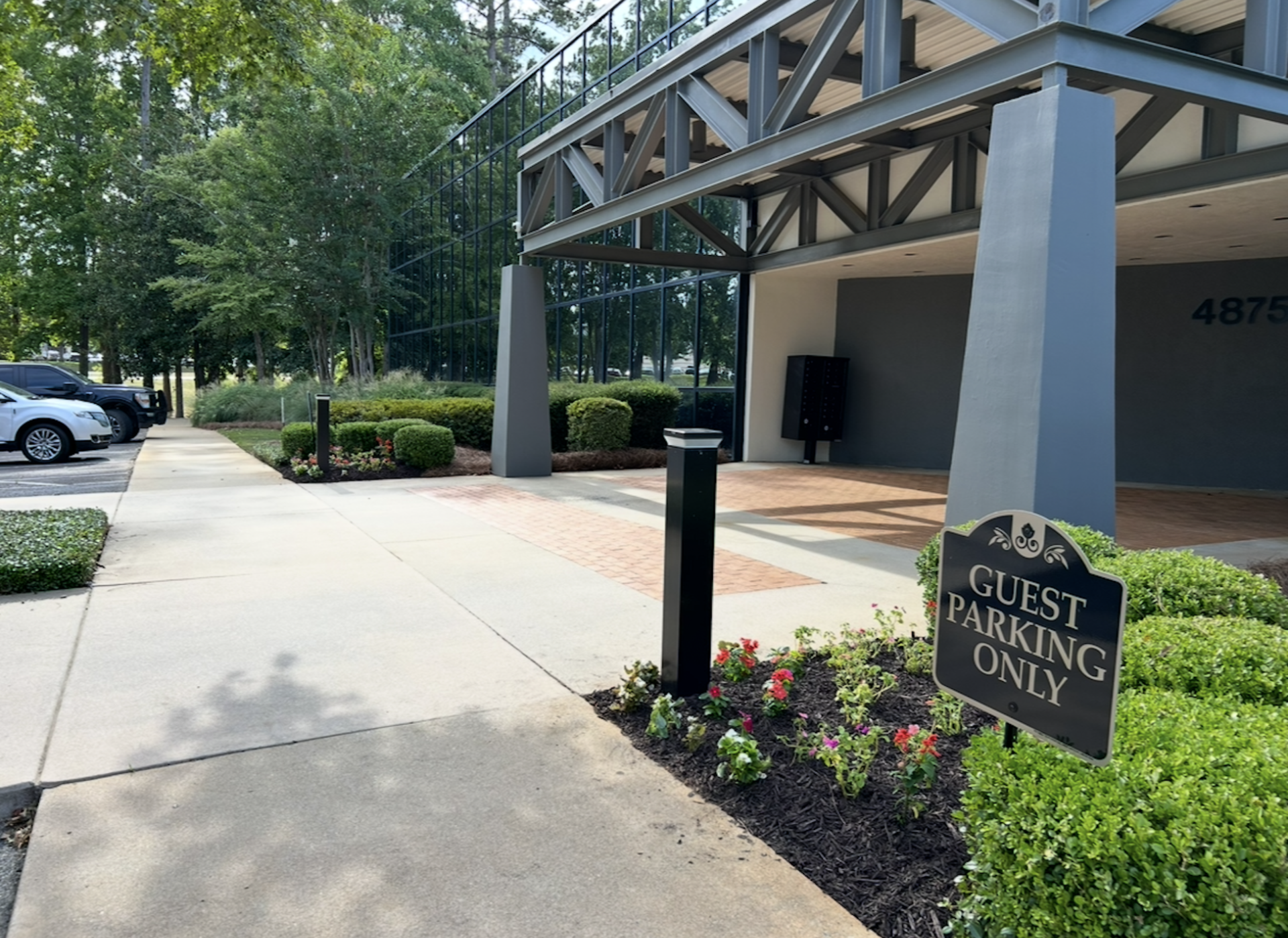 Outdoor walkway beside a modern building with trees, shrubs, and a parked car in the background