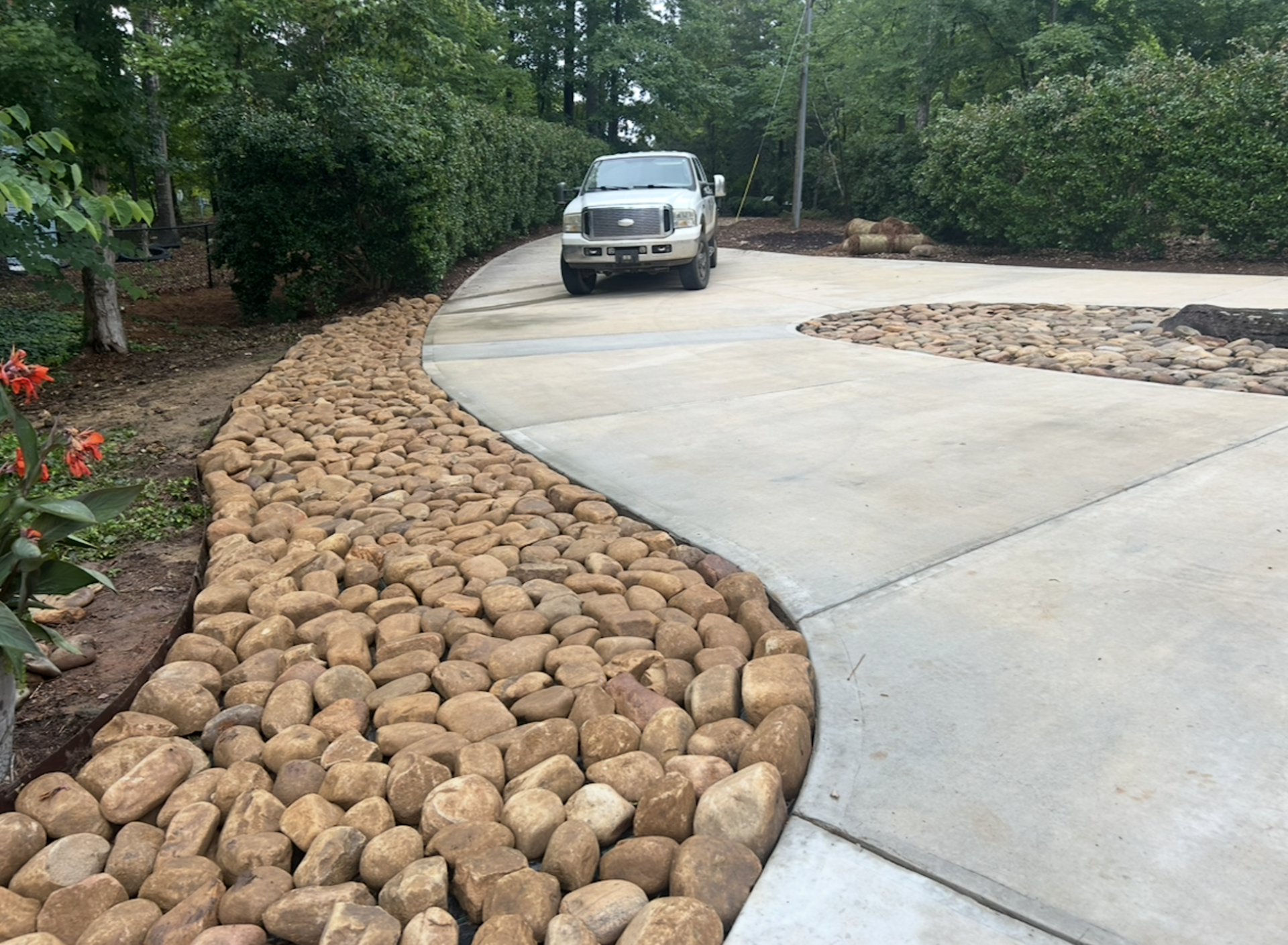 Curved concrete driveway beside a stone-lined garden path with trees and a parked SUV ahead