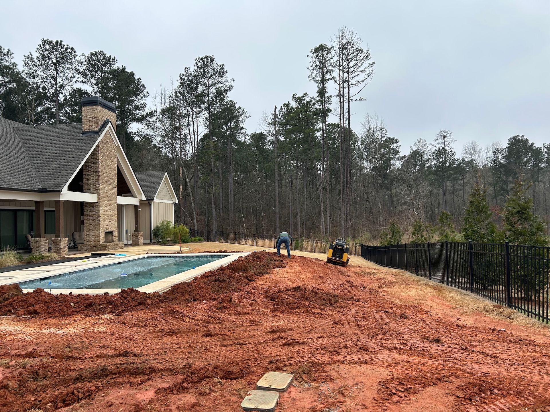 New home construction site with red dirt, pool area, and workers near a stone house in a wooded yard